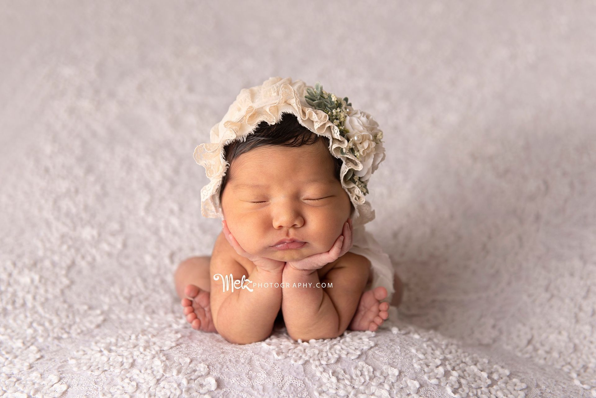 A newborn baby girl wearing a flower headband is sleeping on a white blanket.
