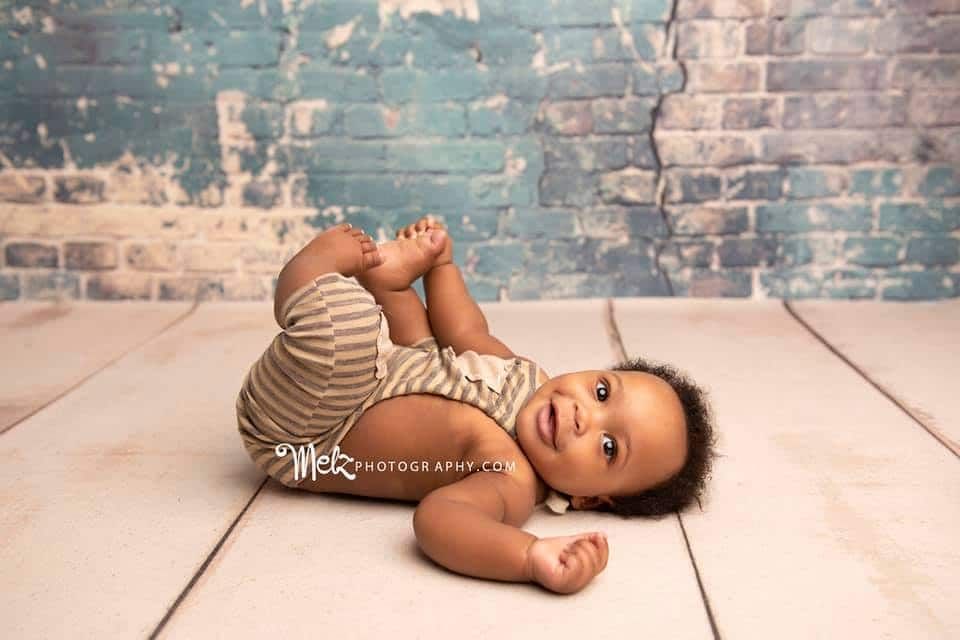 A baby is laying on its stomach on a wooden floor in front of a brick wall.