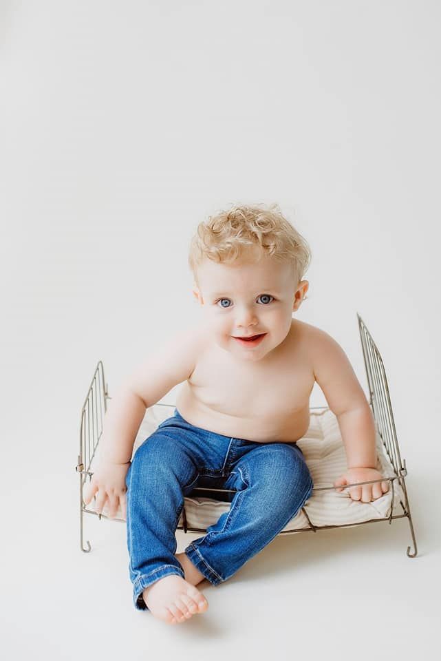 A baby boy without a shirt is sitting on a small bed.