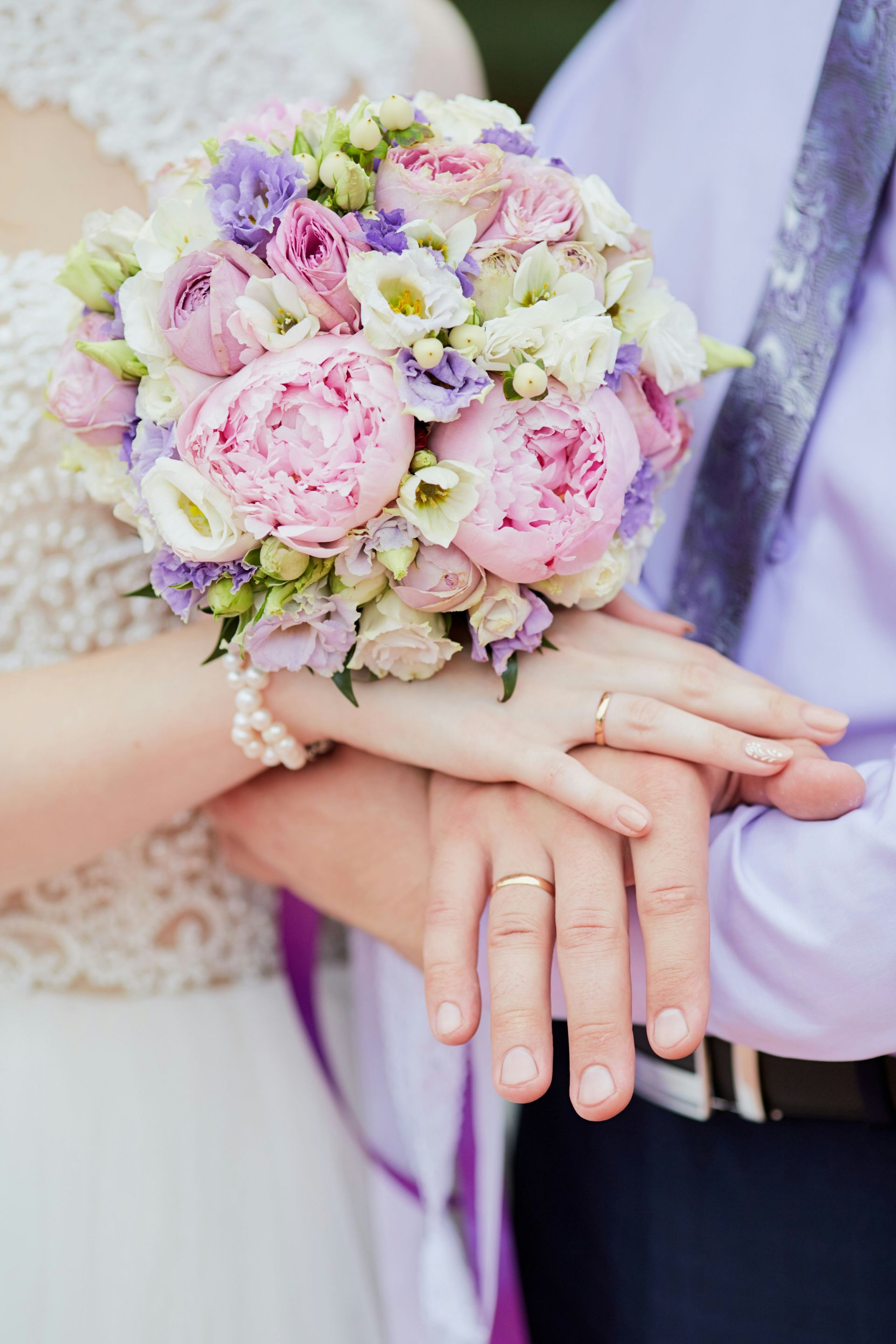 A bride and groom are putting their hands together and holding a bouquet of flowers.