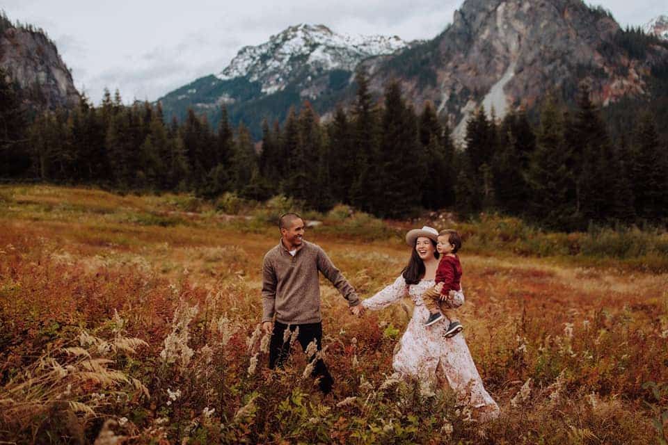 A family is holding hands in a field with mountains in the background.