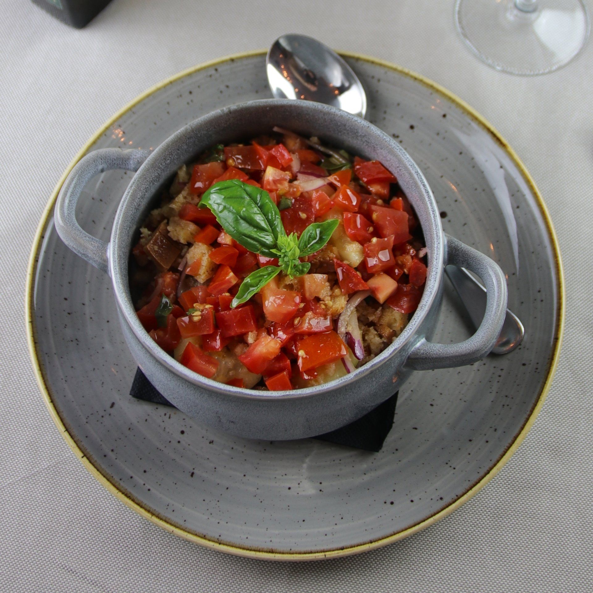 Insalata di pomodori e pane in una ciotola con manico grigio su un piatto, con cucchiaio e guarnizione di basilico.