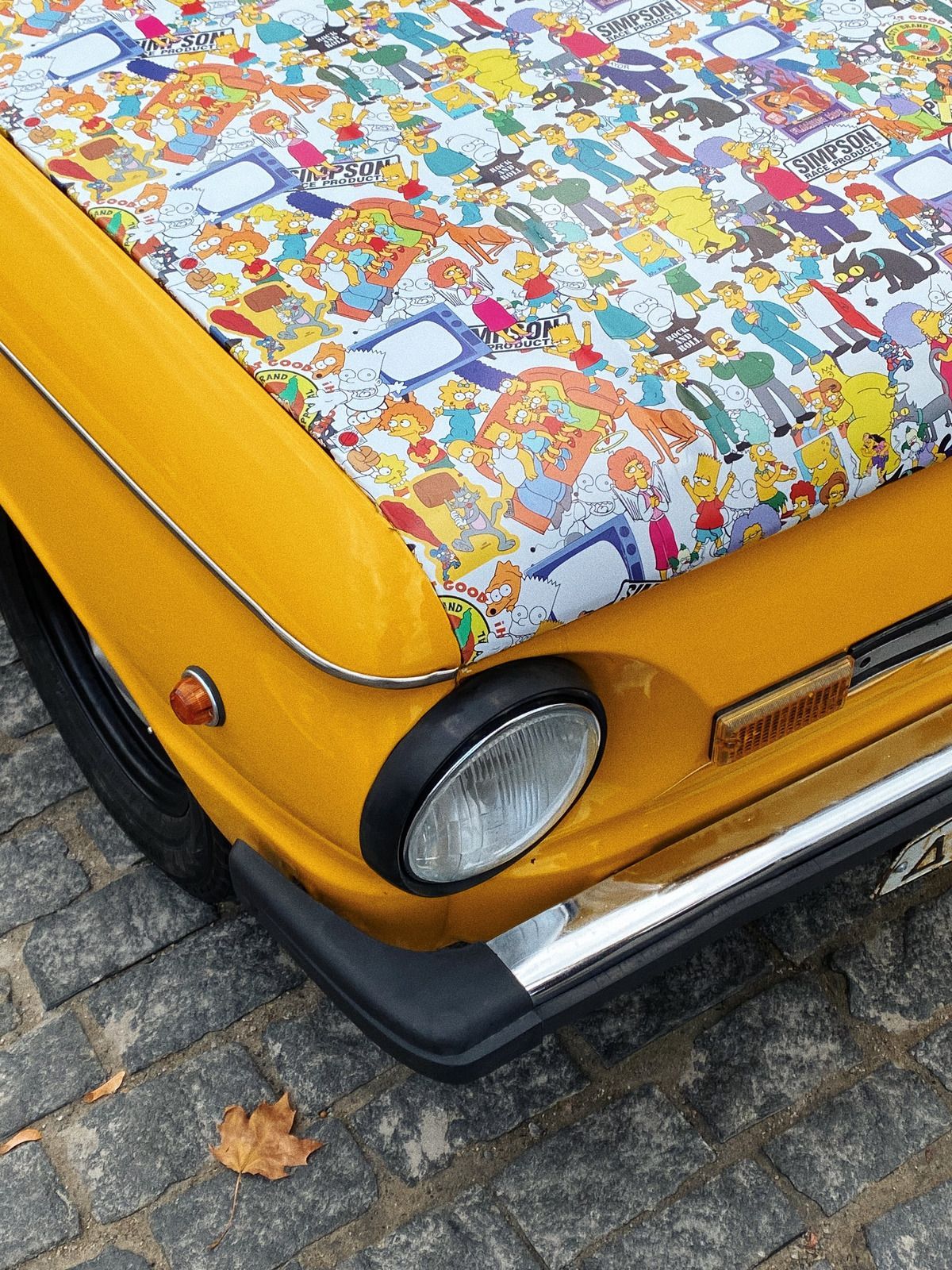 Yellow car hood covered in The Simpsons characters, parked on cobblestone.
