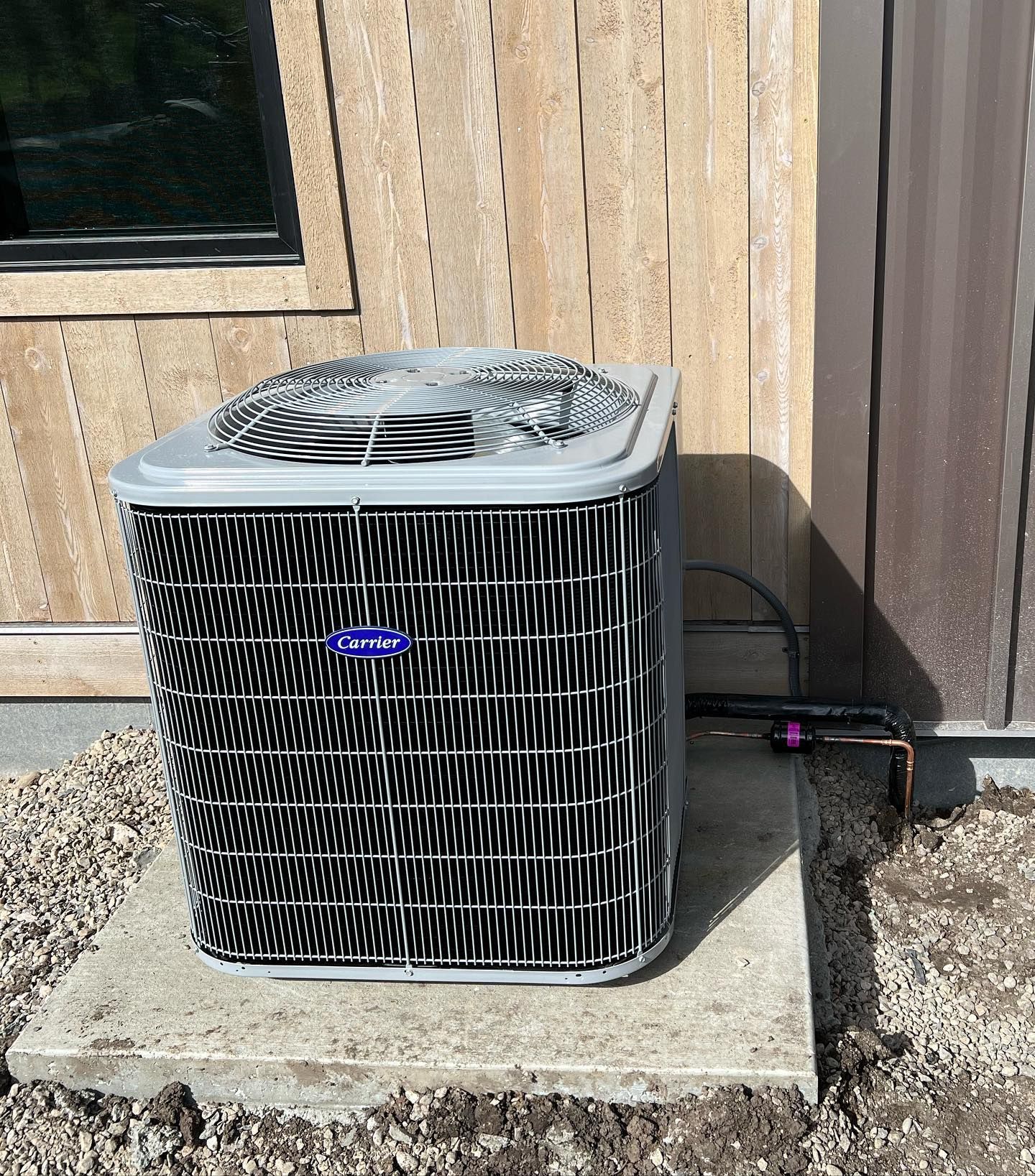 Outdoor air conditioner unit beside a tan wooden house wall on a concrete pad.
