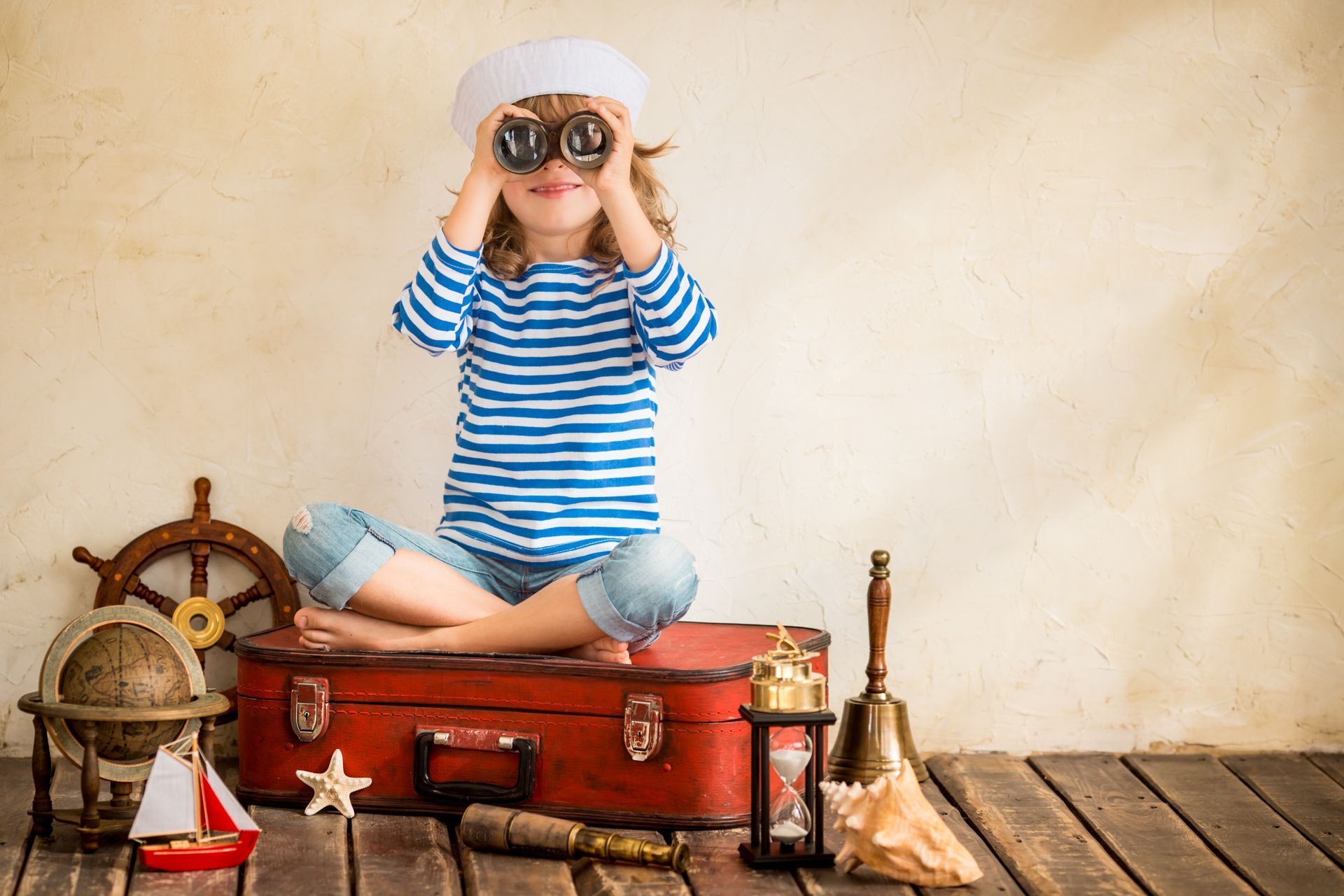 A little girl is sitting on a suitcase looking through binoculars.