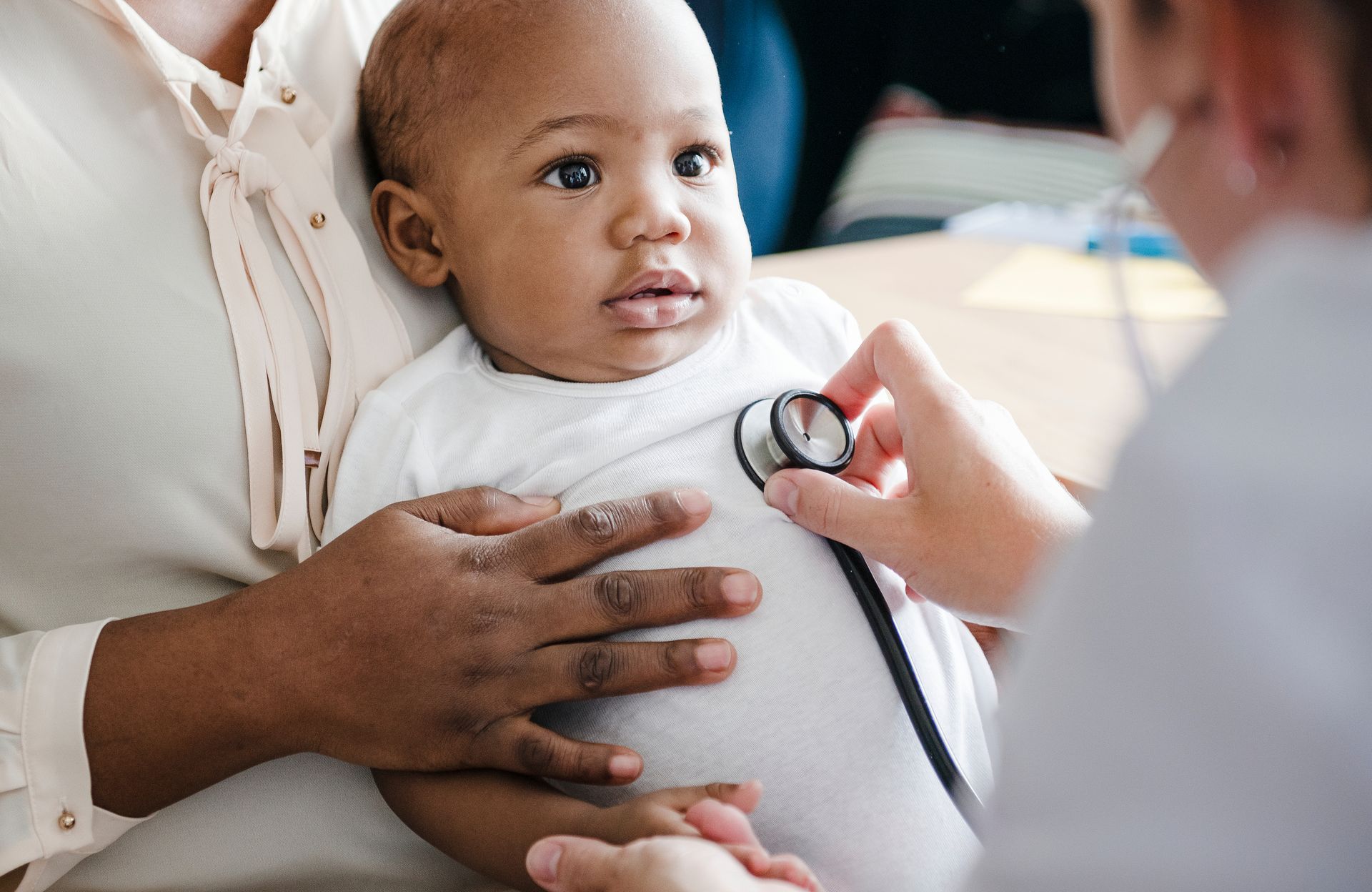 Baby being examined by a doctor with a stethoscope while held by an adult in a clinic