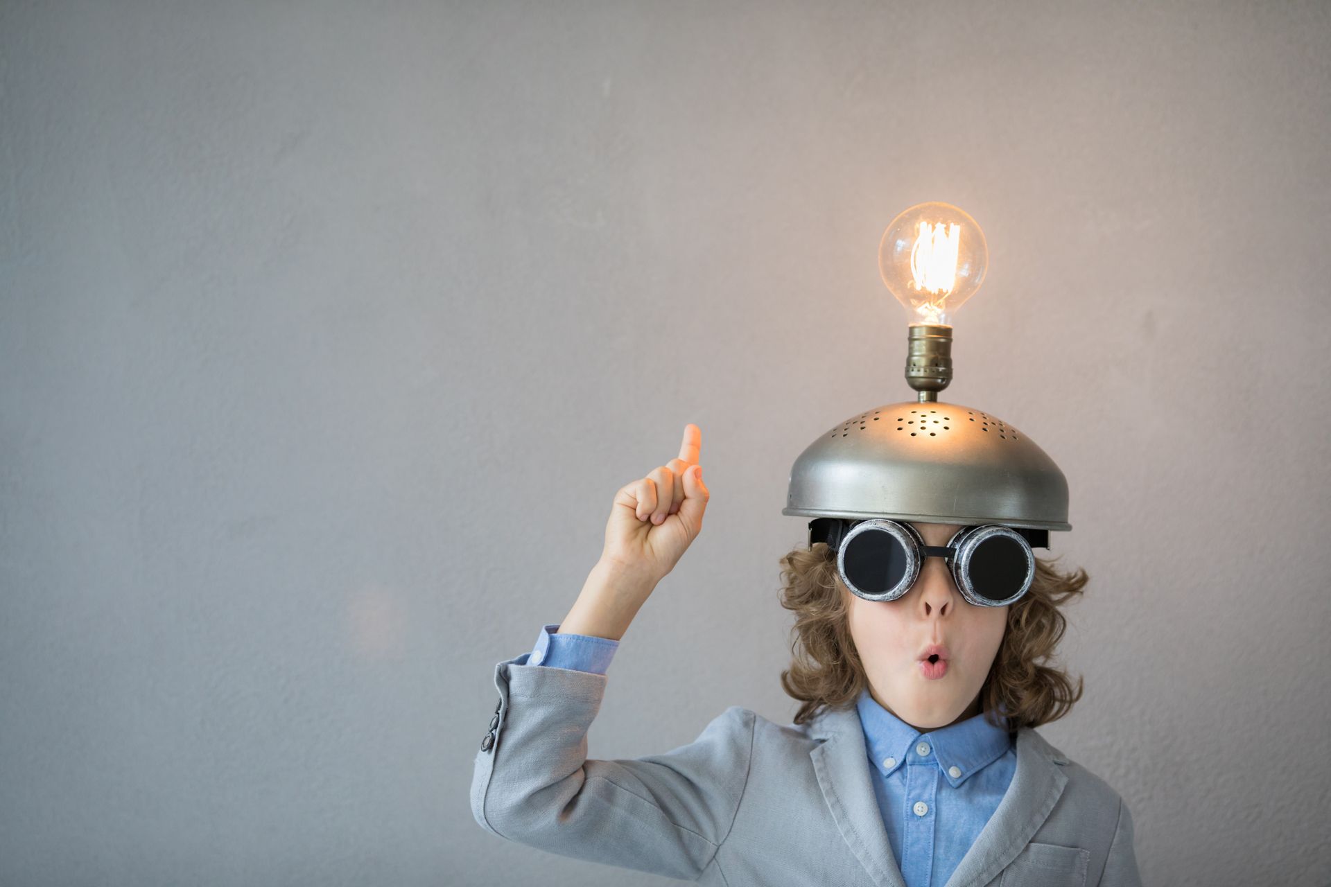 Boy in goggles and helmet with lightbulb on head, pointing up, surprised expression.