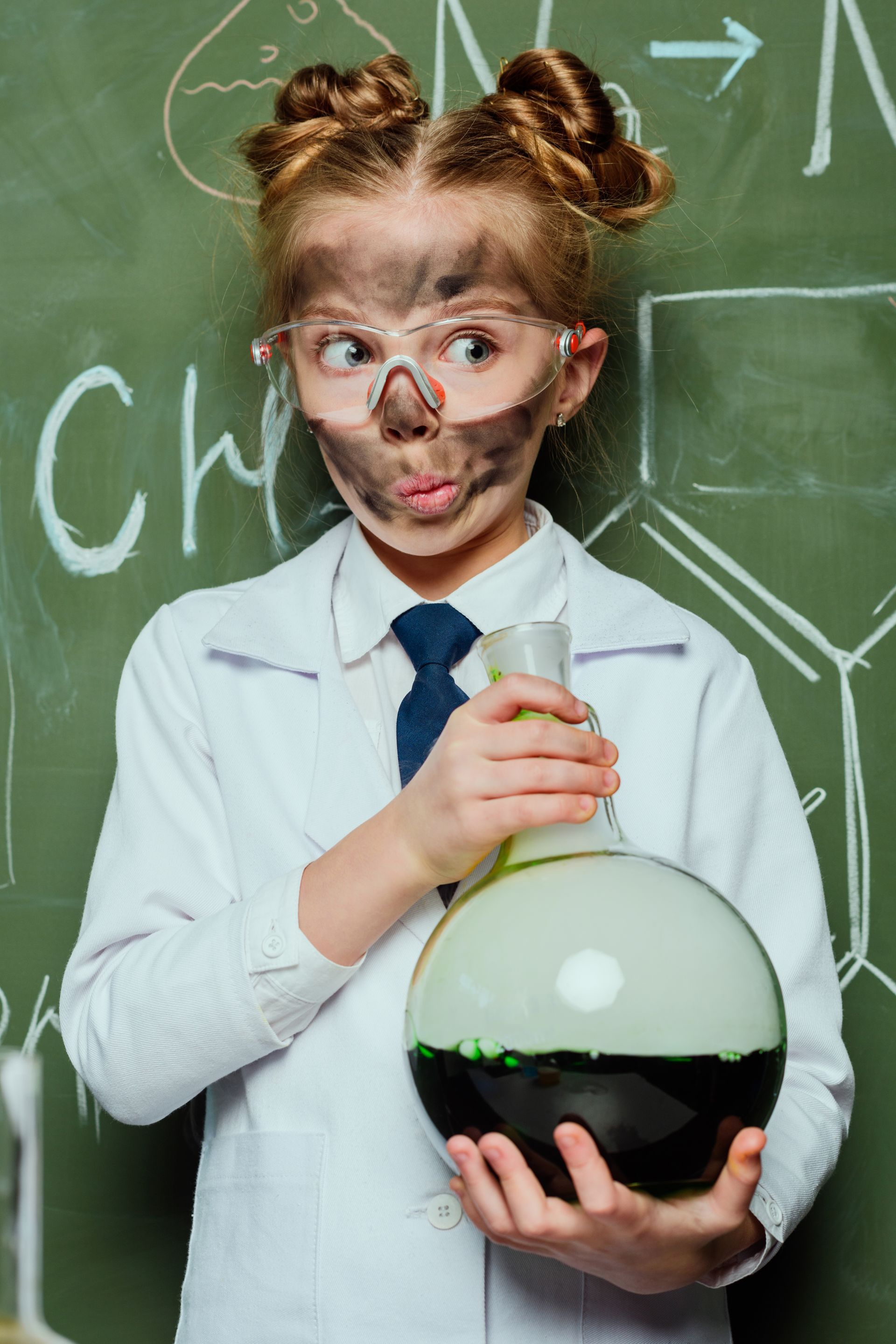 A little girl with a dirty face is holding a beaker in front of a blackboard.