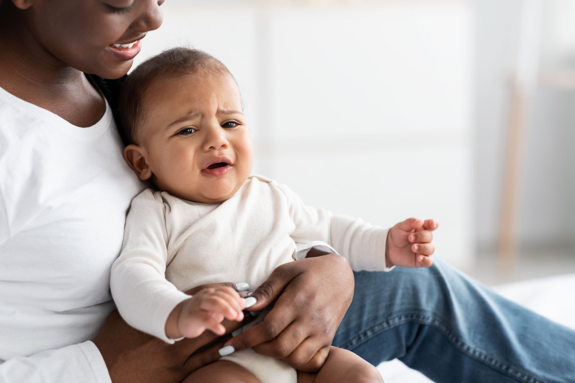 Adult holding a crying baby in a bright living room, both in light clothing.