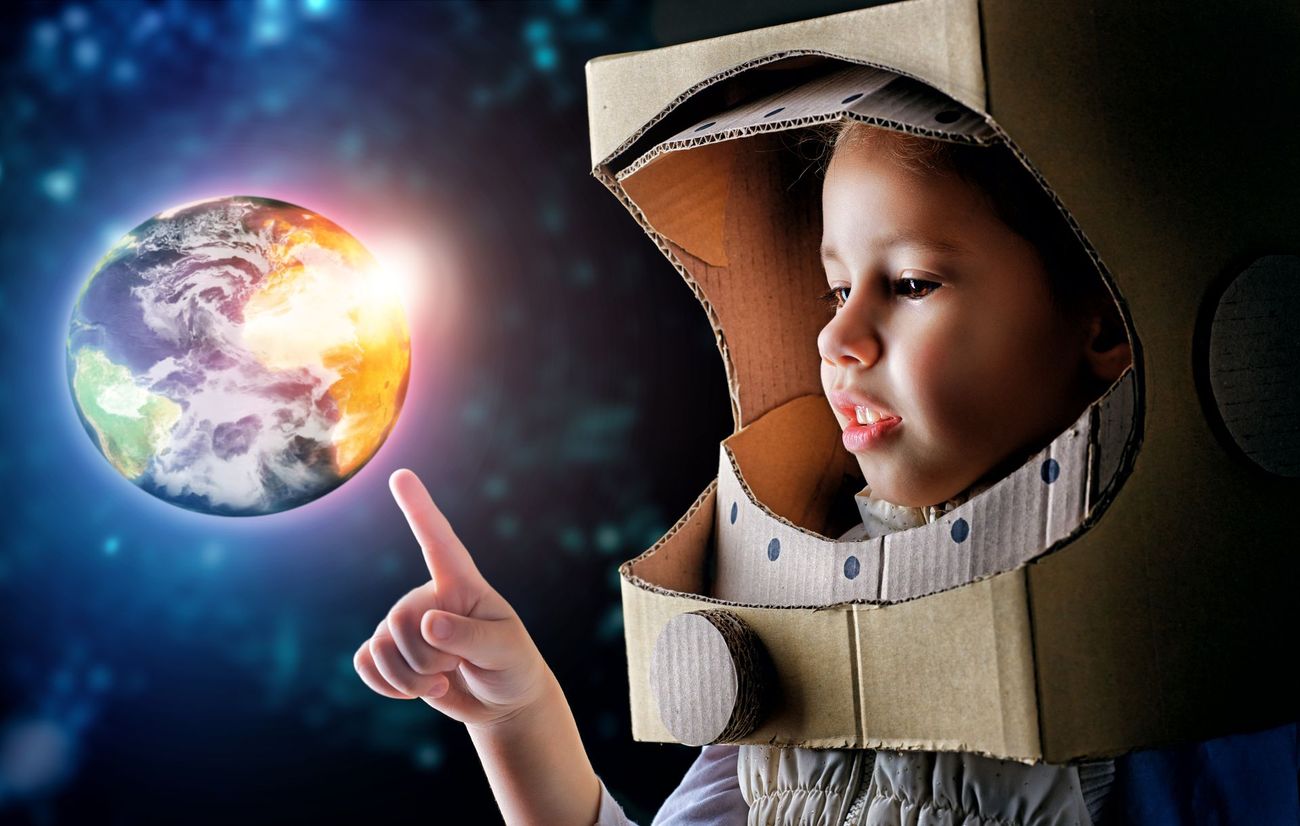 A child is wearing a cardboard space helmet and pointing at the earth.