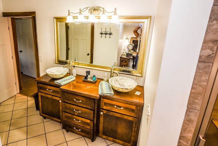 Bathroom with double sinks, gold mirror, wooden cabinets, and a doorway. Steam Shower