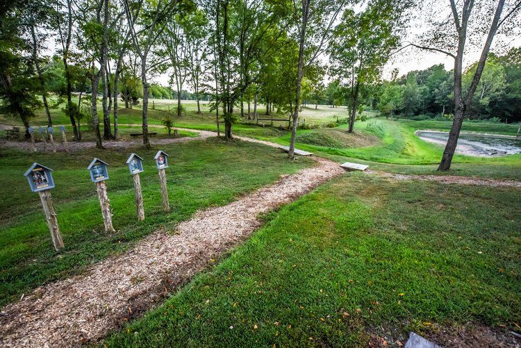 A gravel path leads through a grassy park toward a pond. Birdhouses stand along the prayer path. Trees line the background.