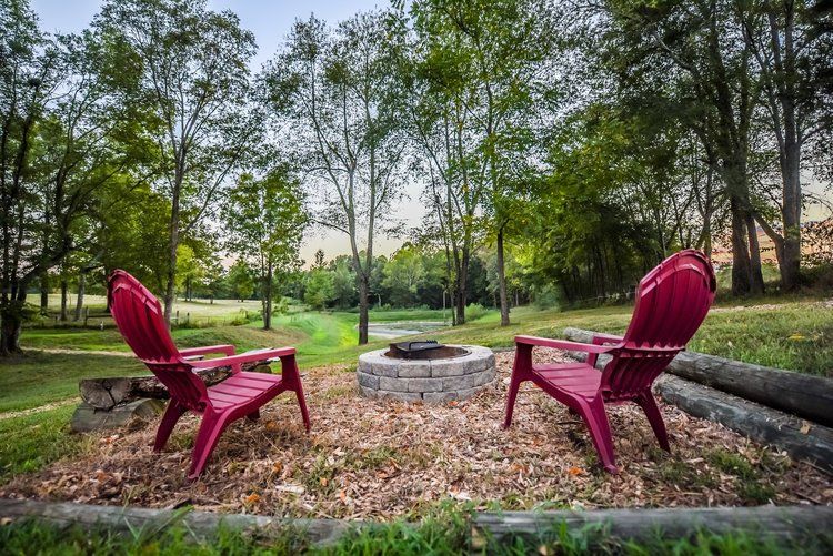 Two red Adirondack chairs face a stone fire pit in a wooded setting.