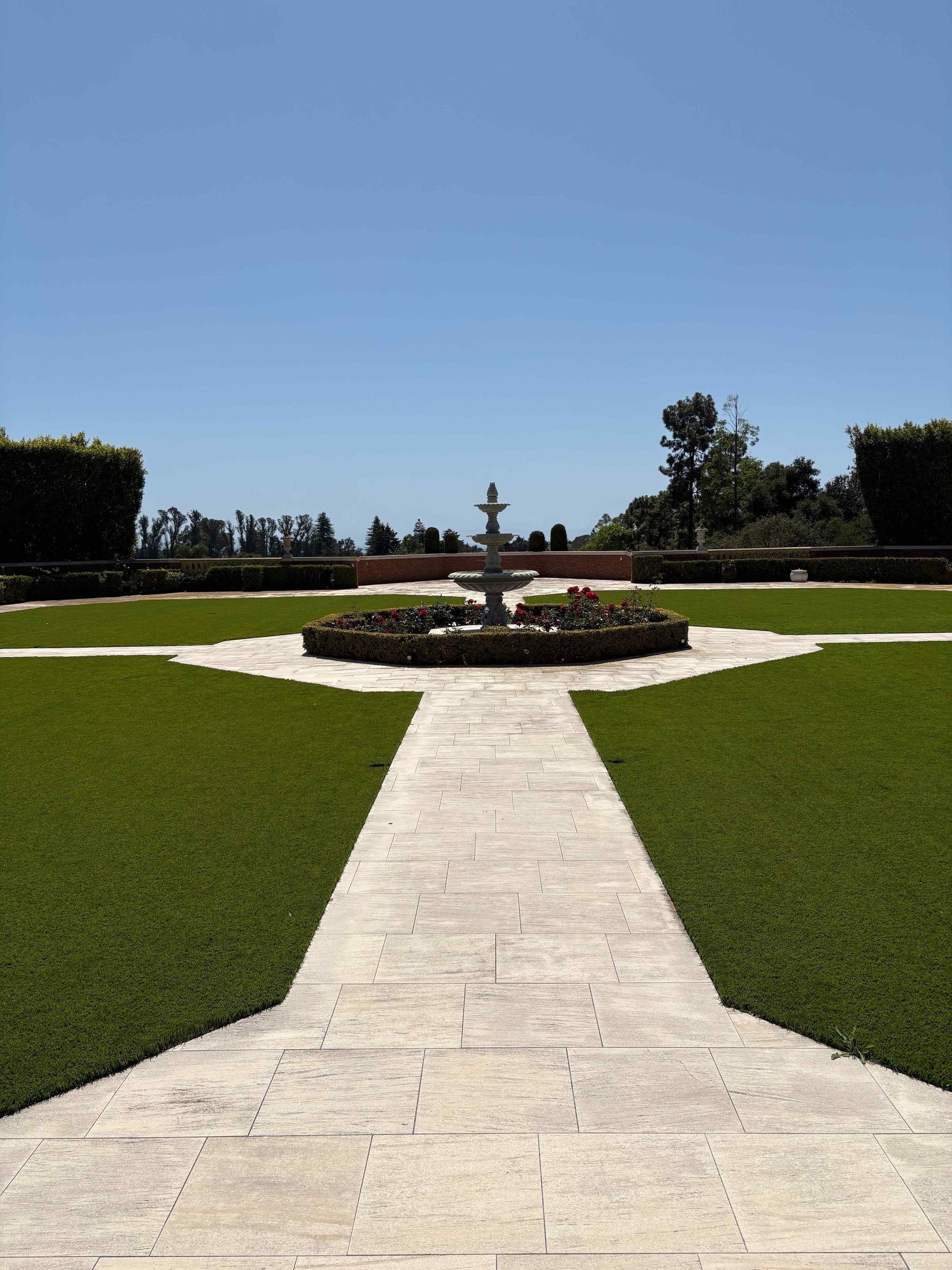 Stone path through green lawn leading to a central fountain in a manicured garden under a blue sky