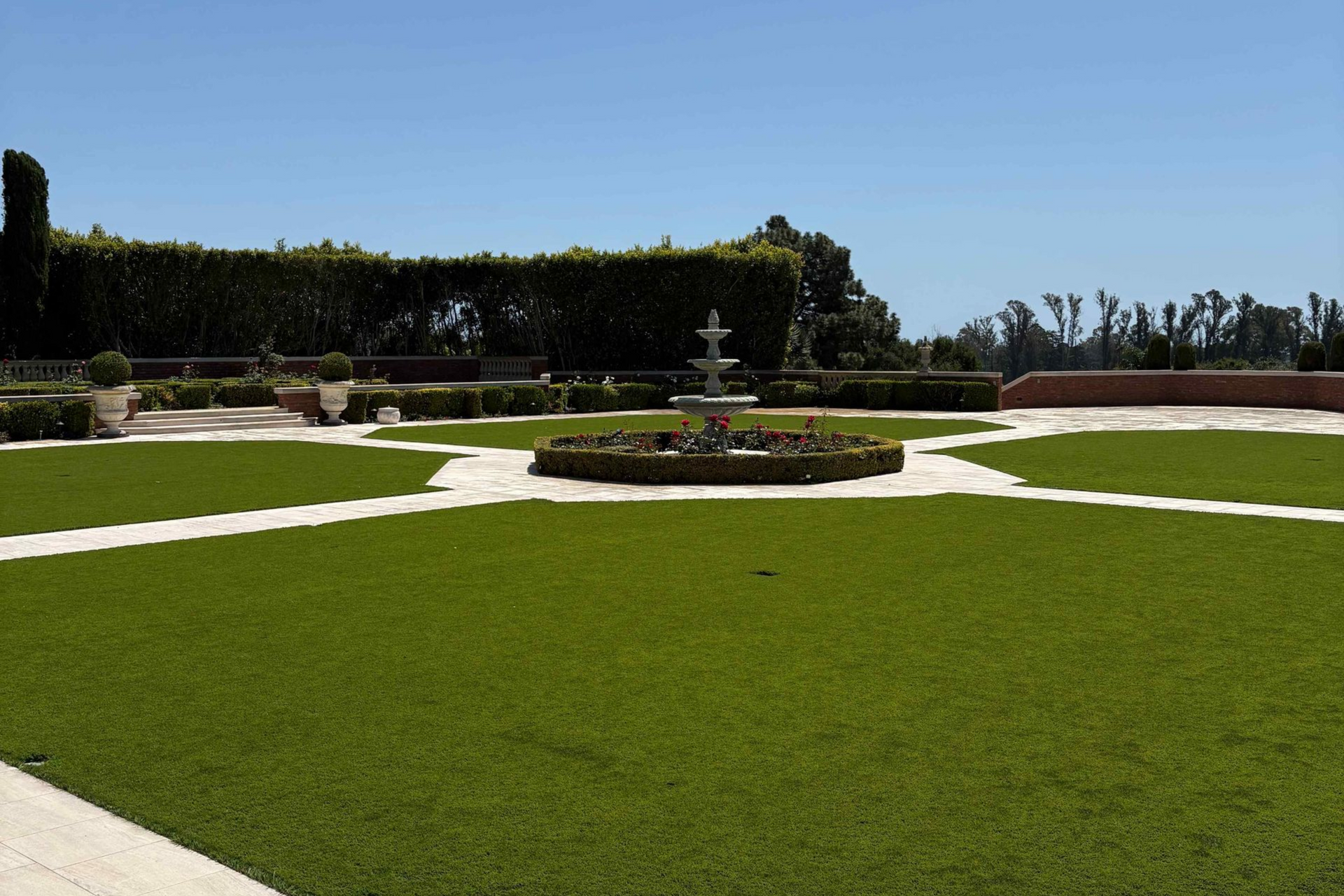 Formal garden with a circular fountain, green lawn, white paths, and hedges under a clear blue sky