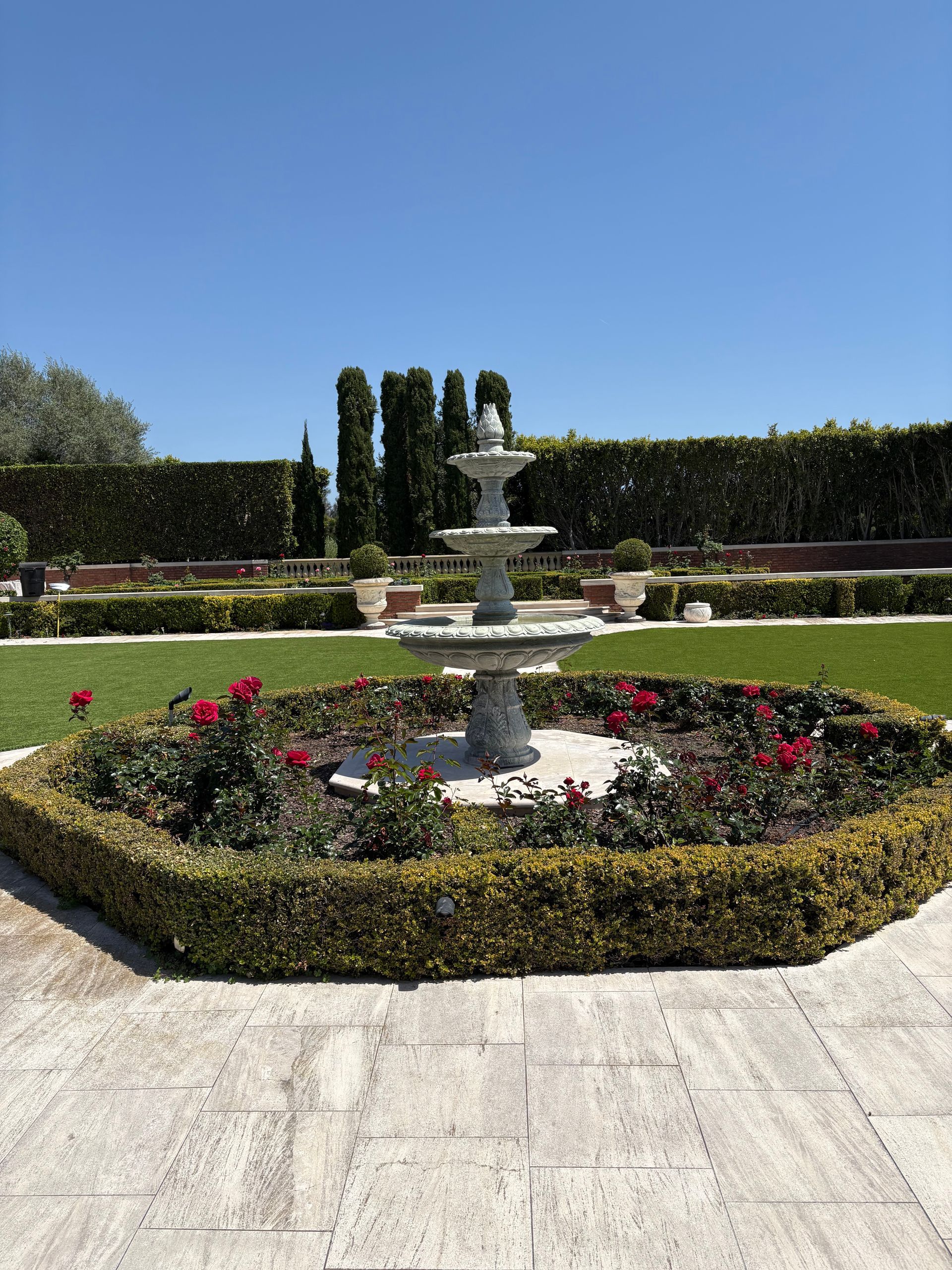 Stone fountain in a formal garden, surrounded by red flowers and trimmed hedges under a clear blue sky