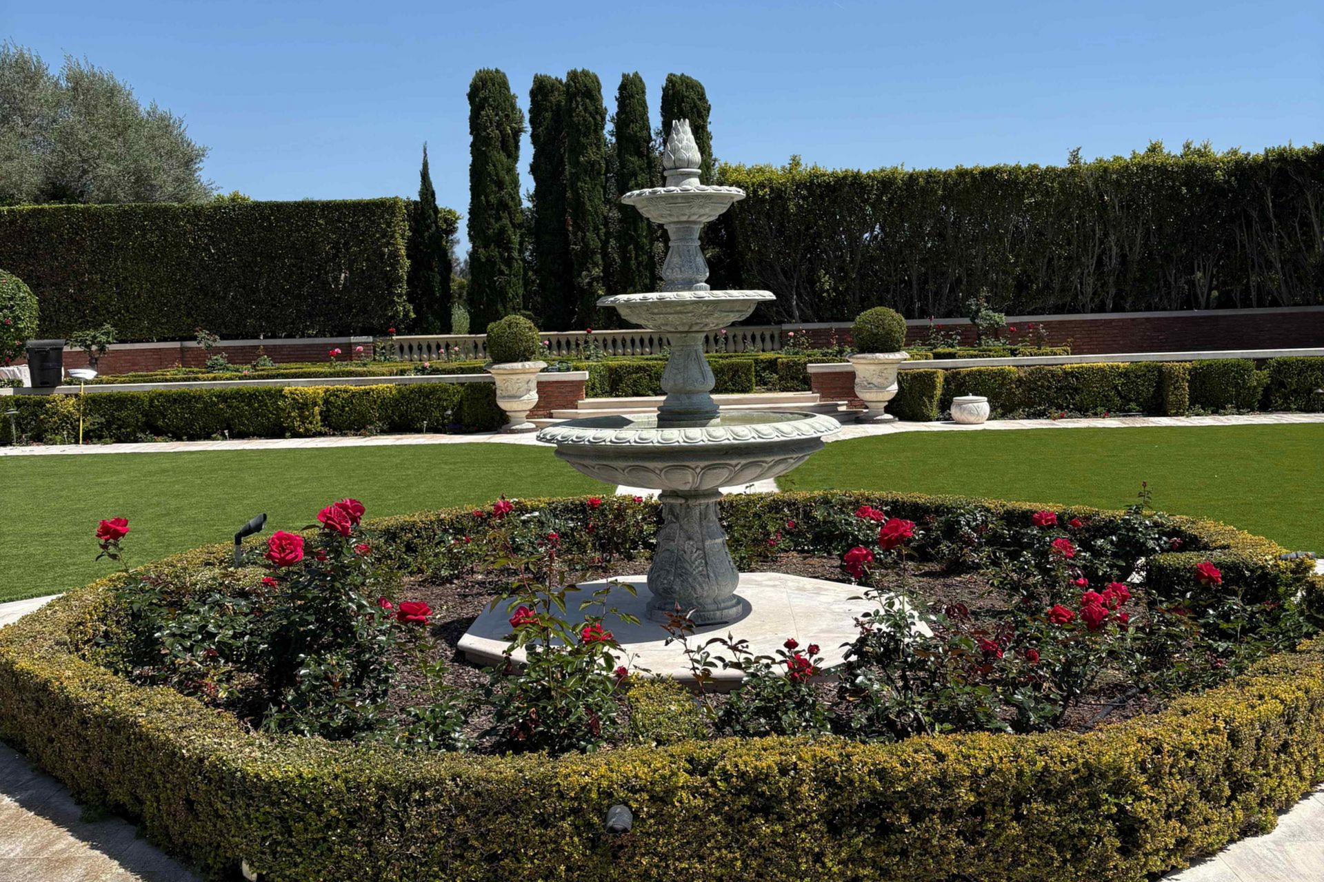 Tiered stone fountain in a manicured garden with red roses and trimmed hedges under a clear sky
