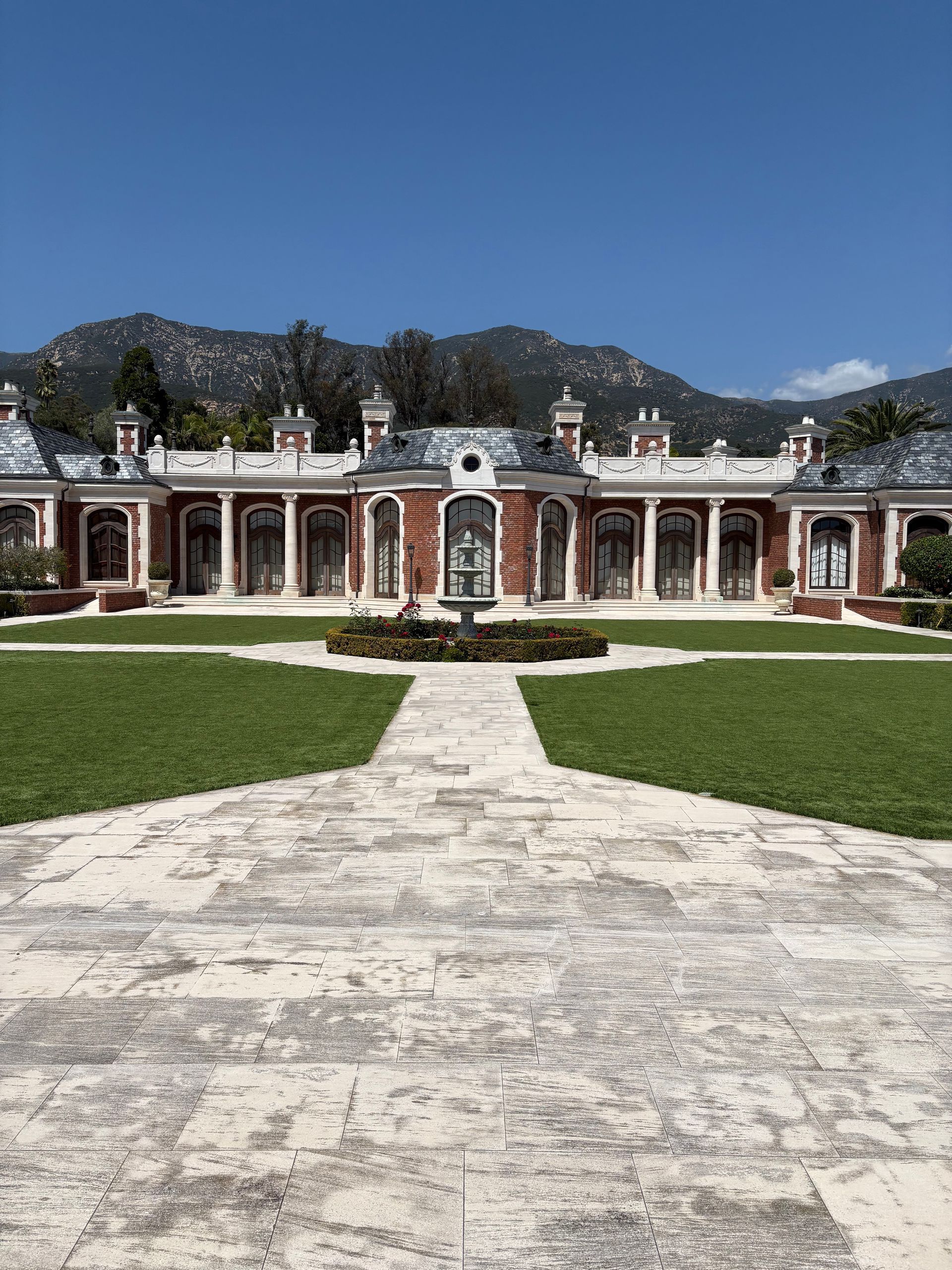 Historic mansion with red columns, centered on a stone path and green lawn, backed by mountains under a blue sky
