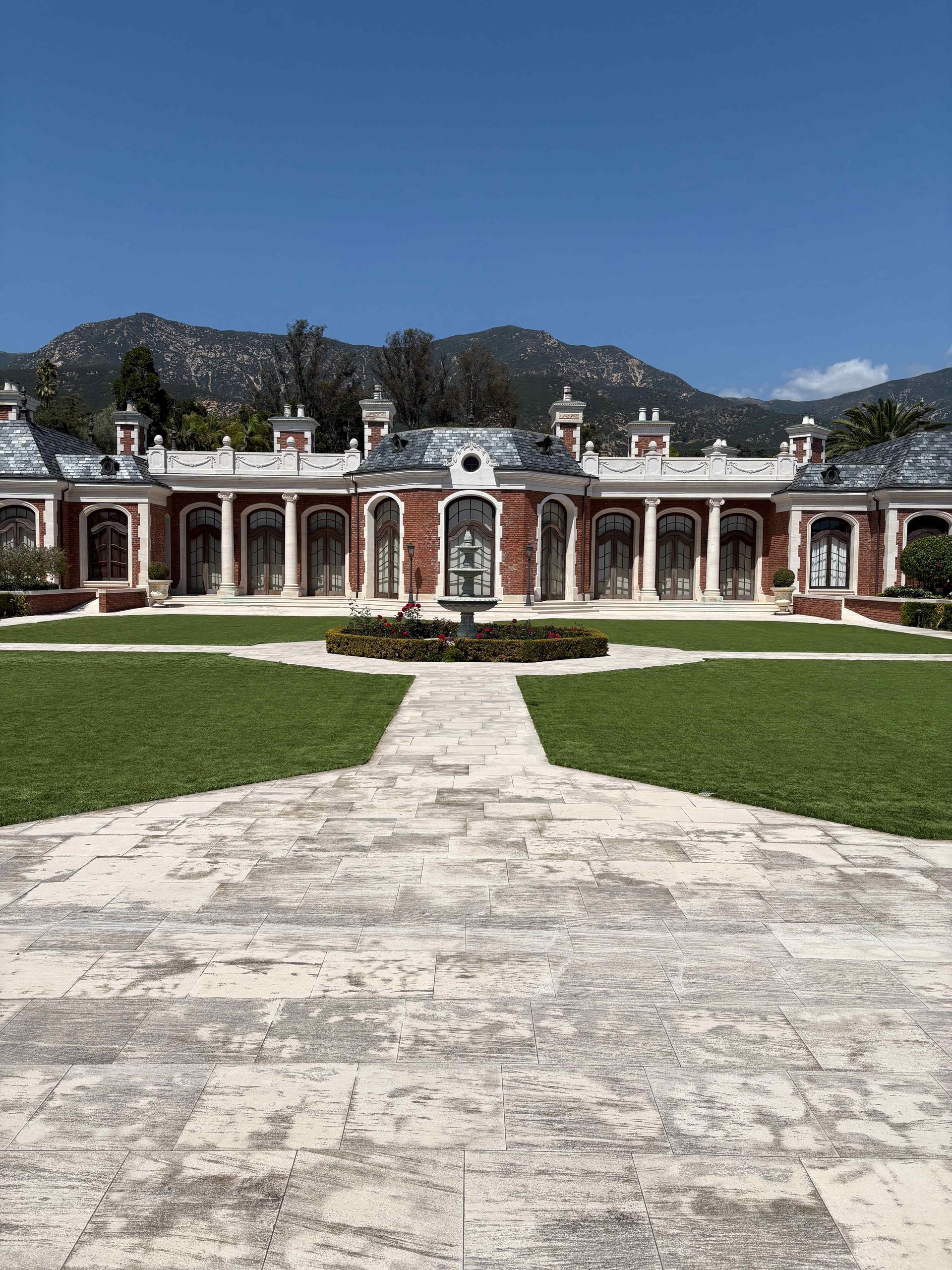Historic red-and-white mansion with a symmetrical facade, central path, and mountains under a clear blue sky.