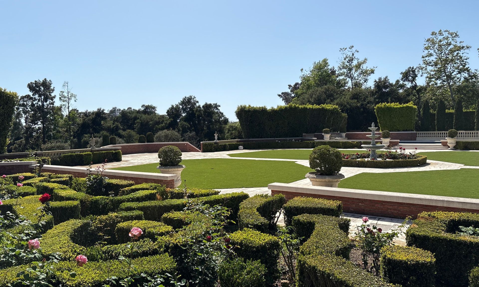 Formal garden with trimmed hedges, brick paths, and a fountain under a clear blue sky.