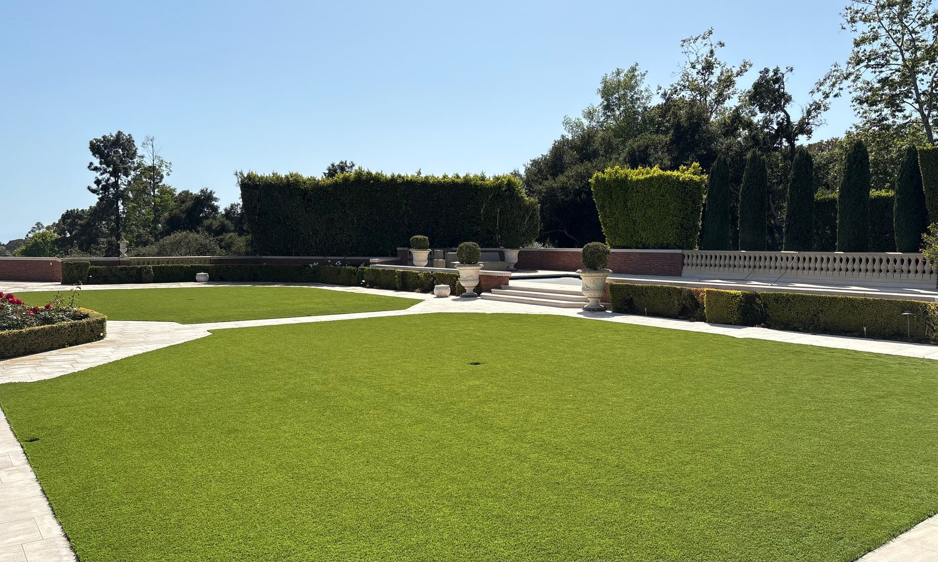 Sunny garden with a green lawn, stone paths, hedges, and flowerbeds under a clear blue sky
