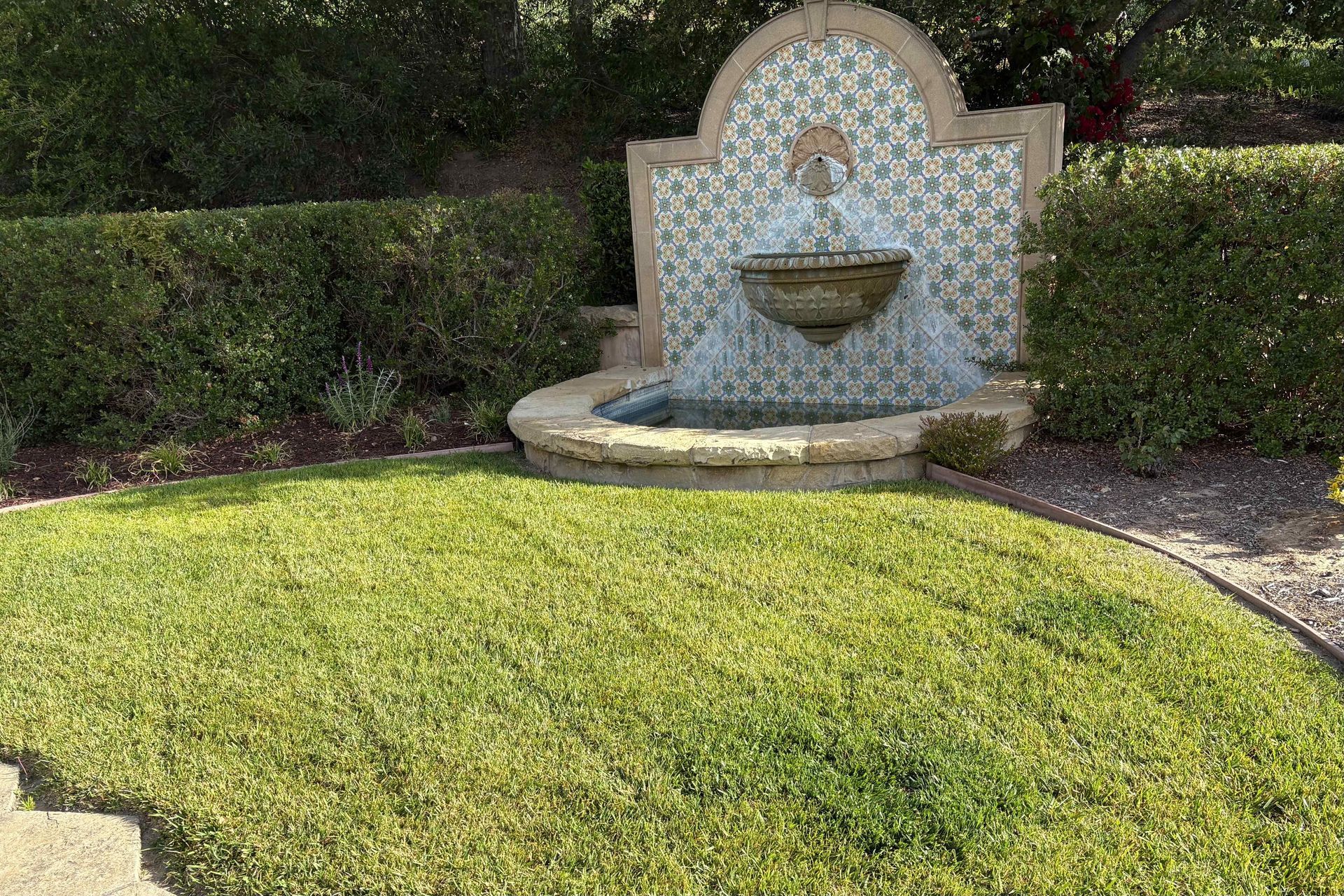 Small decorative fountain with blue mosaic tiles in a landscaped garden beside a green lawn.