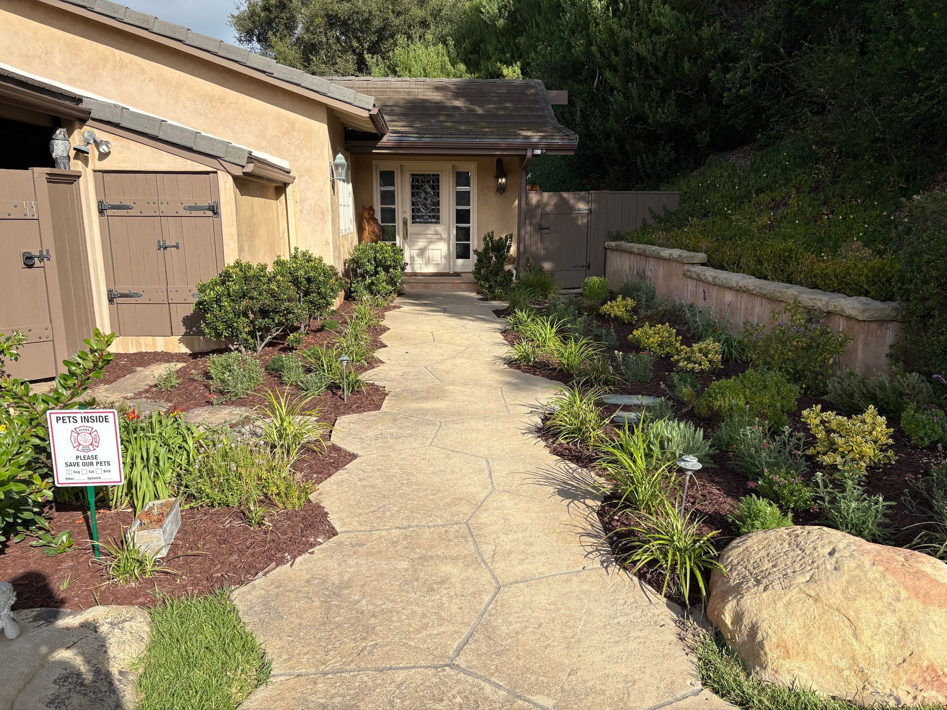 Front walkway to a house with desert landscaping, tan garage, and stone path bordered by shrubs.