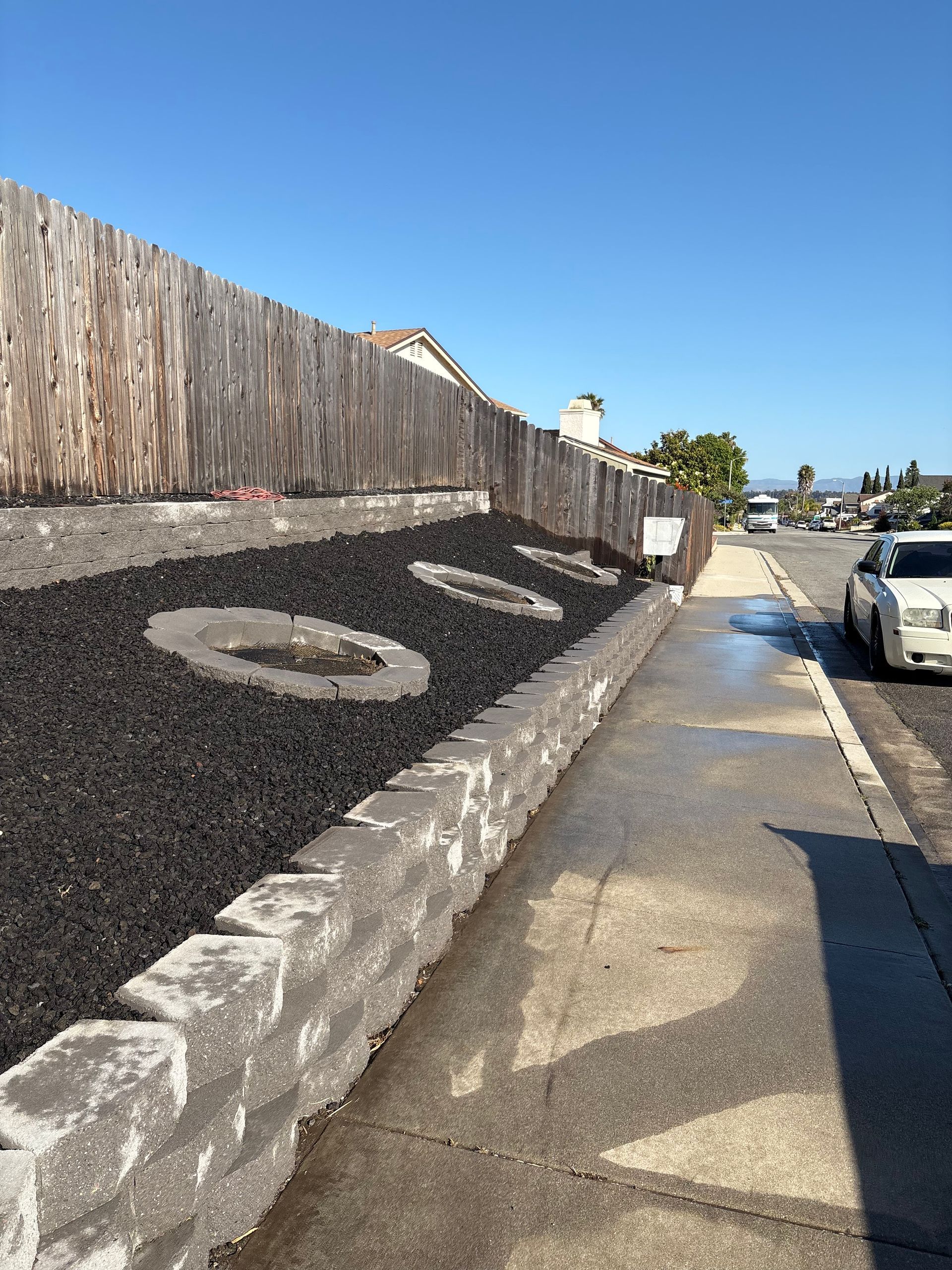 Driveway beside a black mulch landscaped slope with wooden fence and parked cars under a clear blue sky