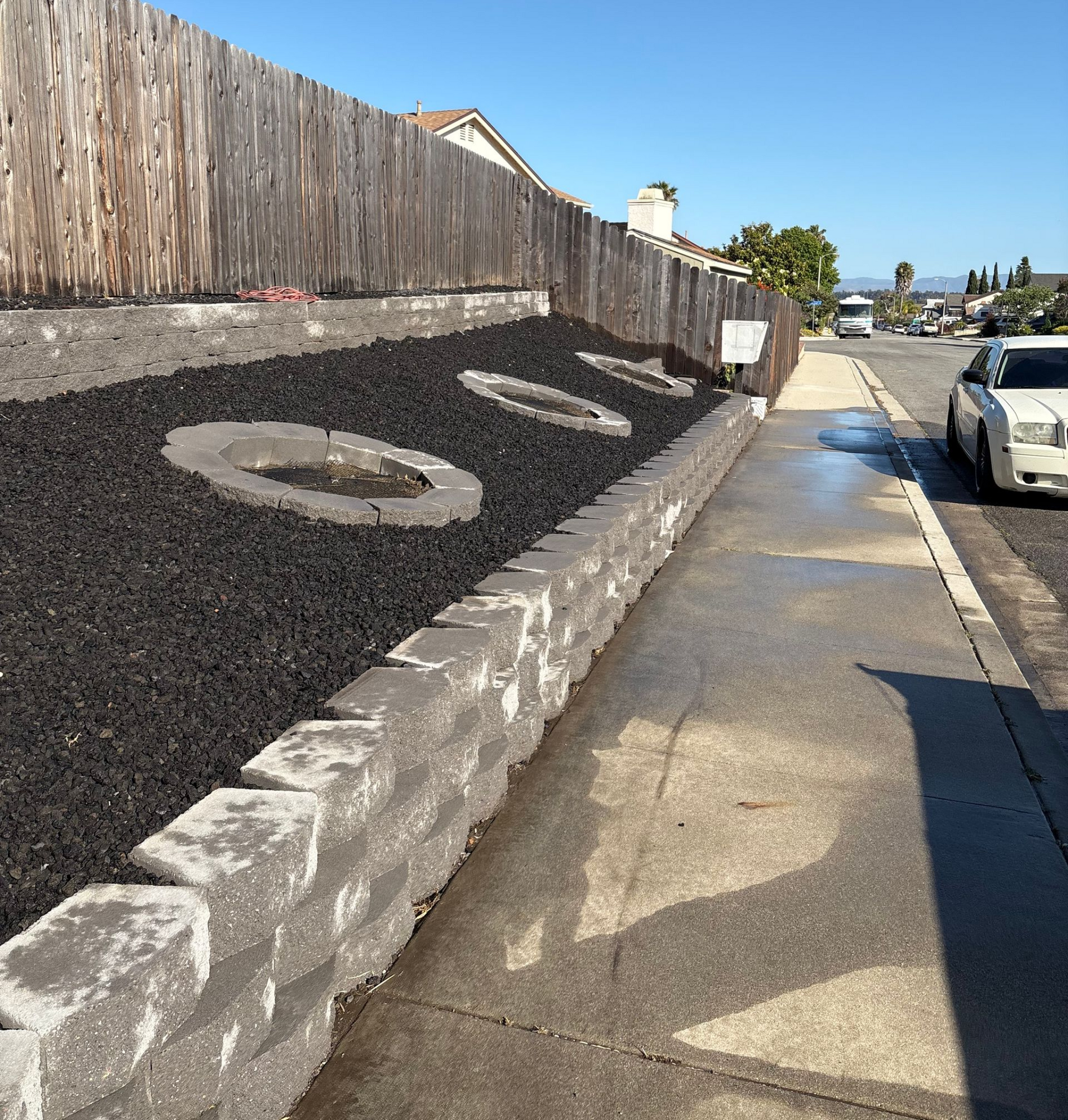 Sloped roadside sign wall with large white letters beside a sidewalk and parked car under blue sky