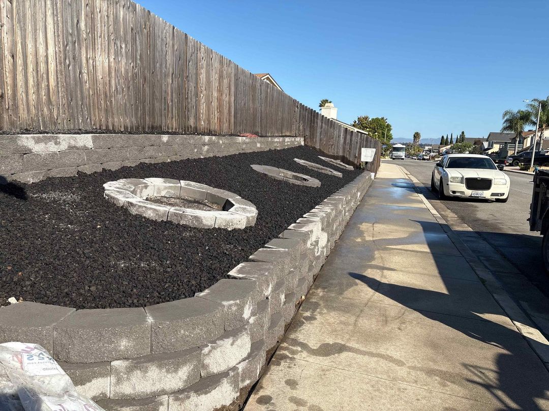 Sloped black planter wall with large white letters beside a sidewalk and parked cars on a sunny street