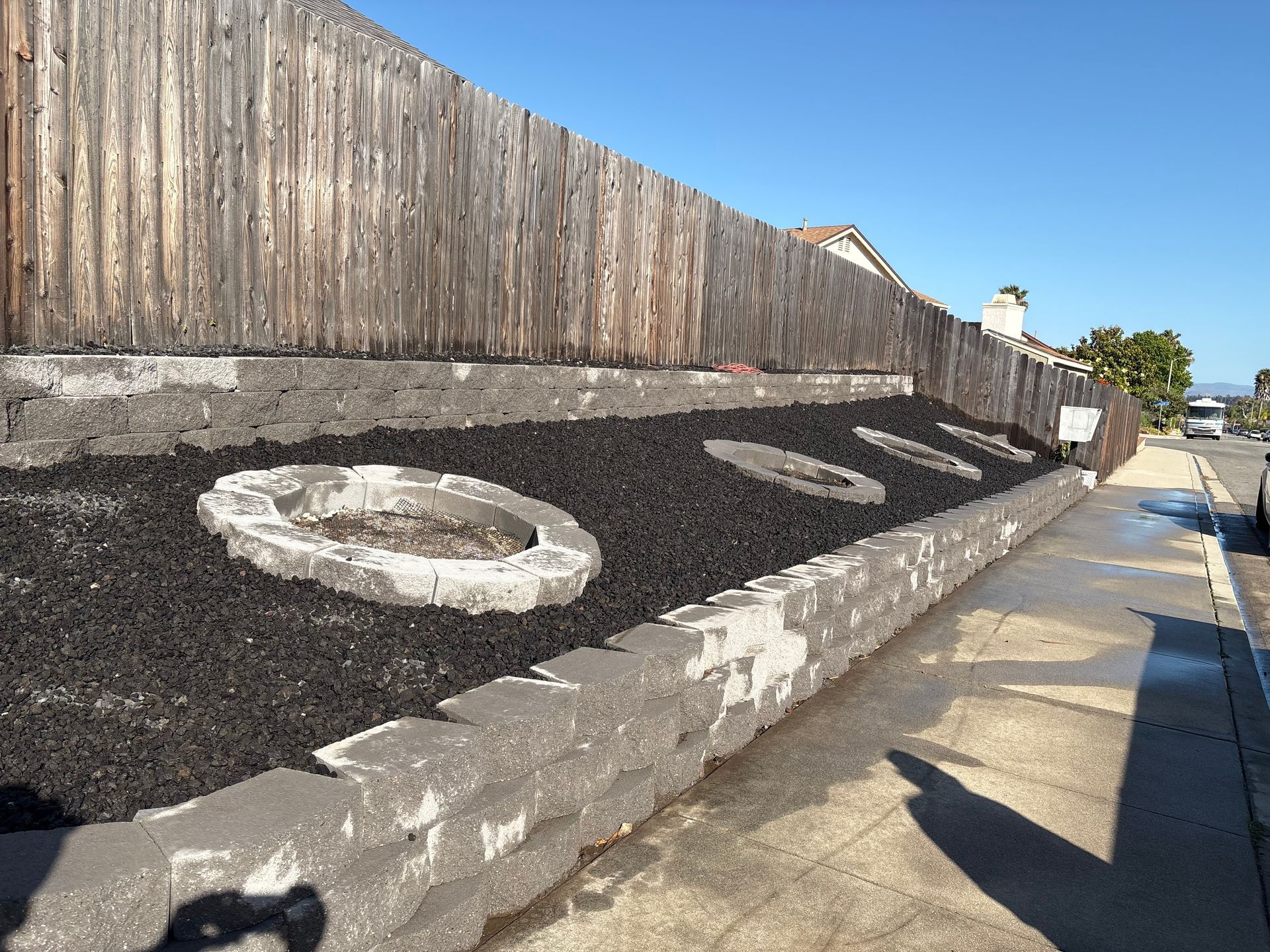Sloped roadside wall with black mulch, white stone circles, and a wooden fence under a clear blue sky