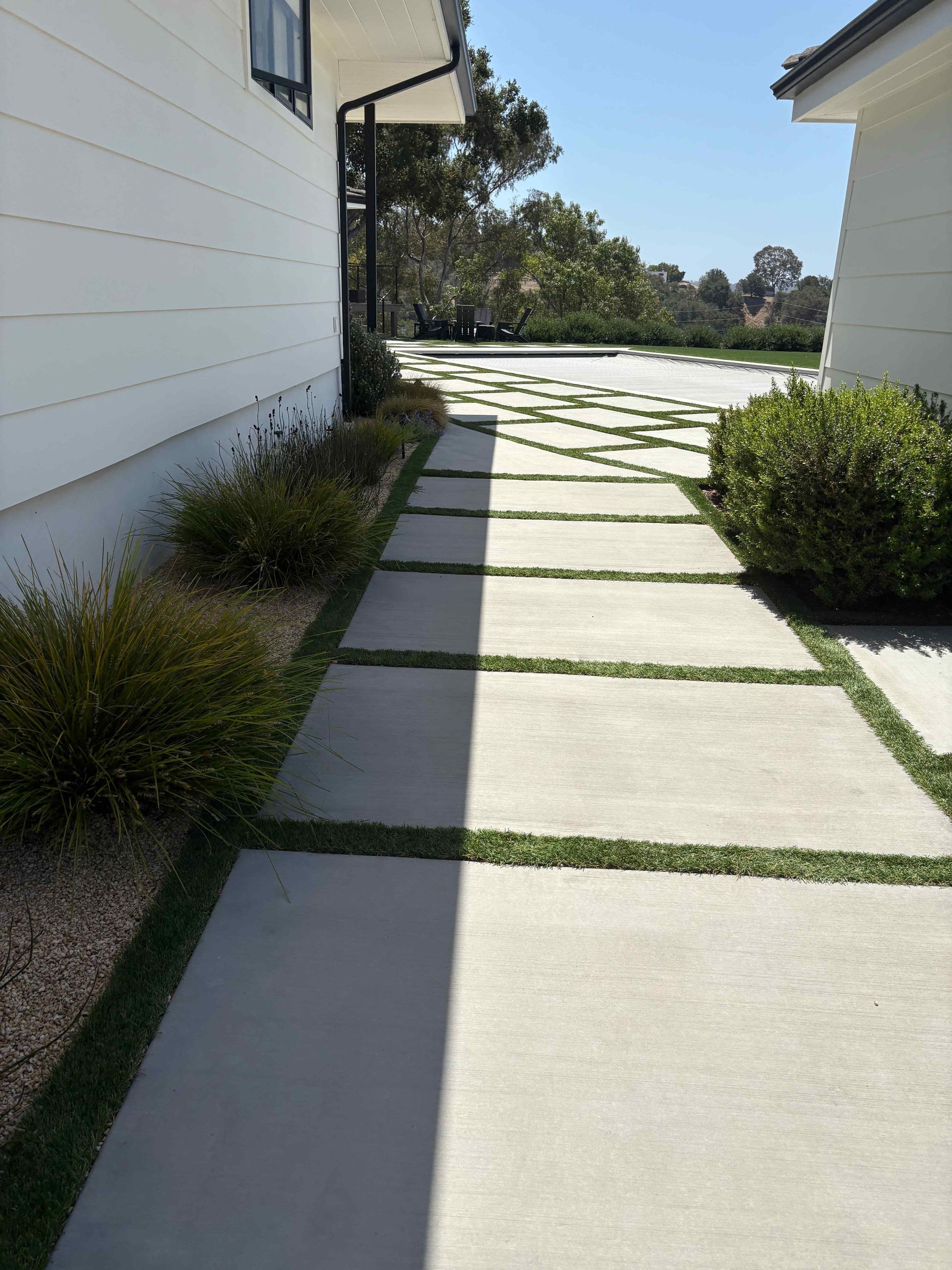 Sunlit concrete path beside a white building, bordered by shrubs and leading to a garden area.