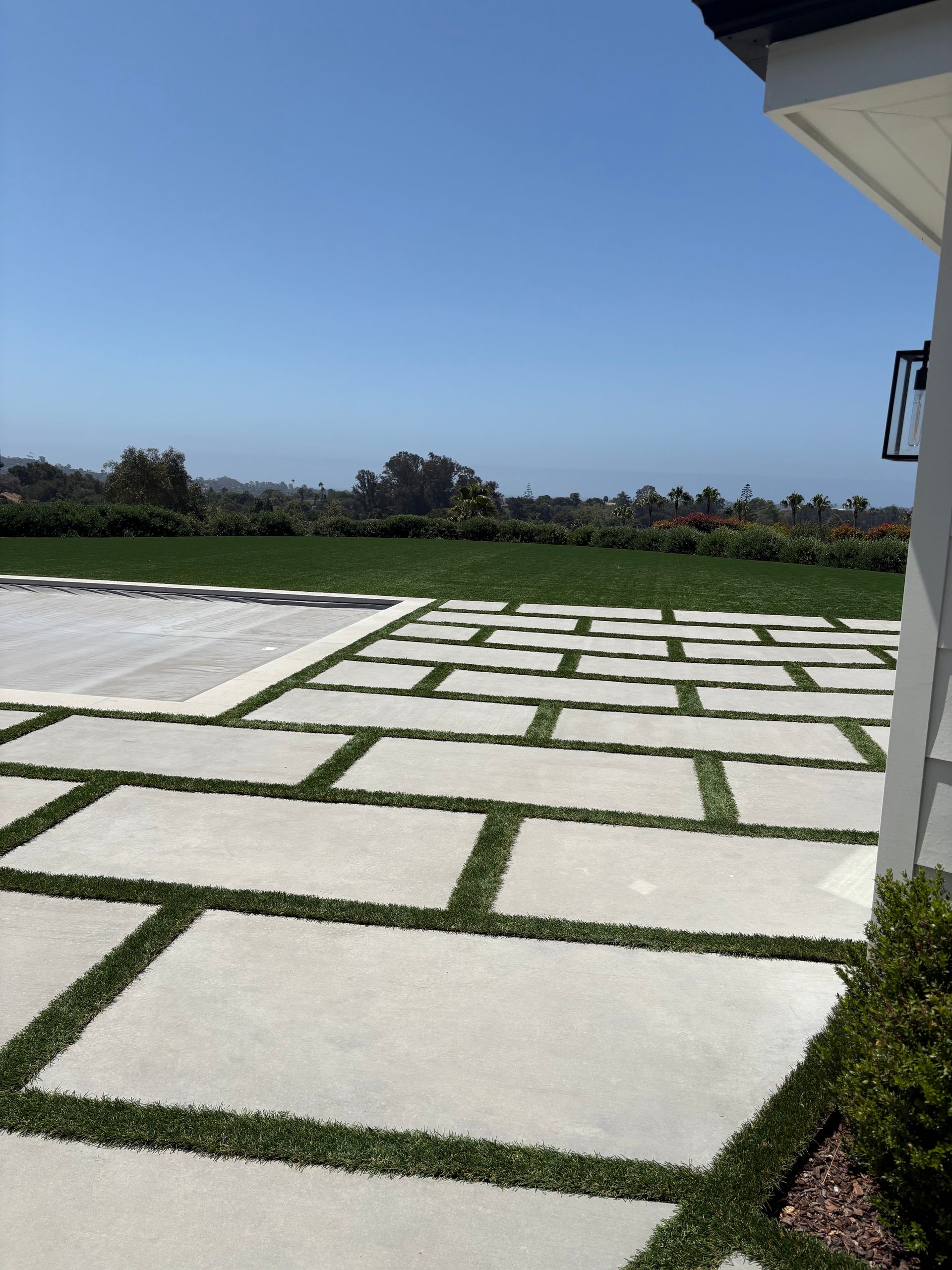 White stone patio with green grass borders, overlooking a grassy field under a clear blue sky