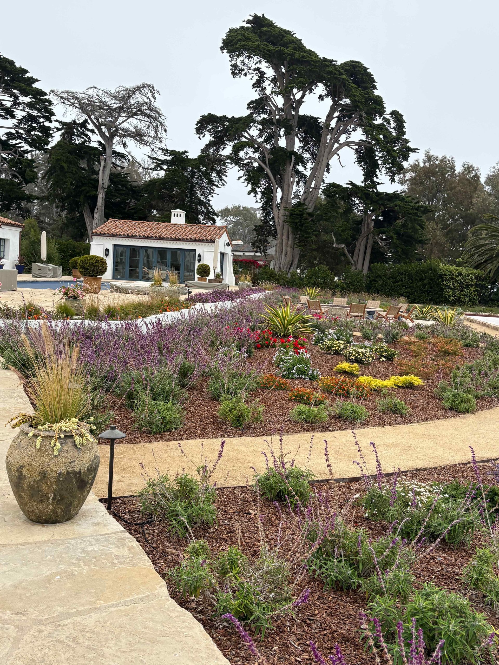 Colorful flower garden with winding paths, a large tree, and a small white building in the background