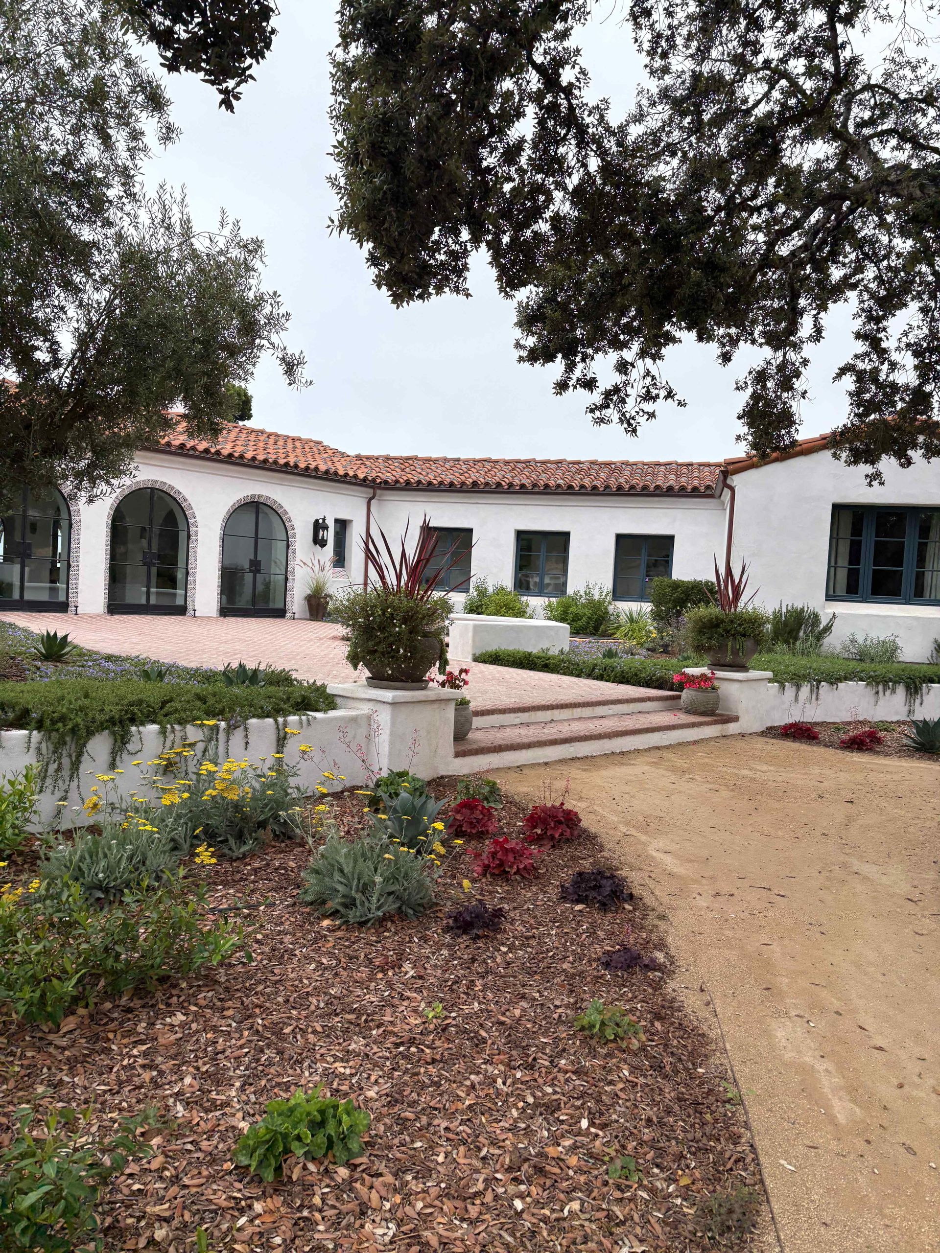 White stucco garden house with red-tile roof, arched windows, and landscaped front path under trees