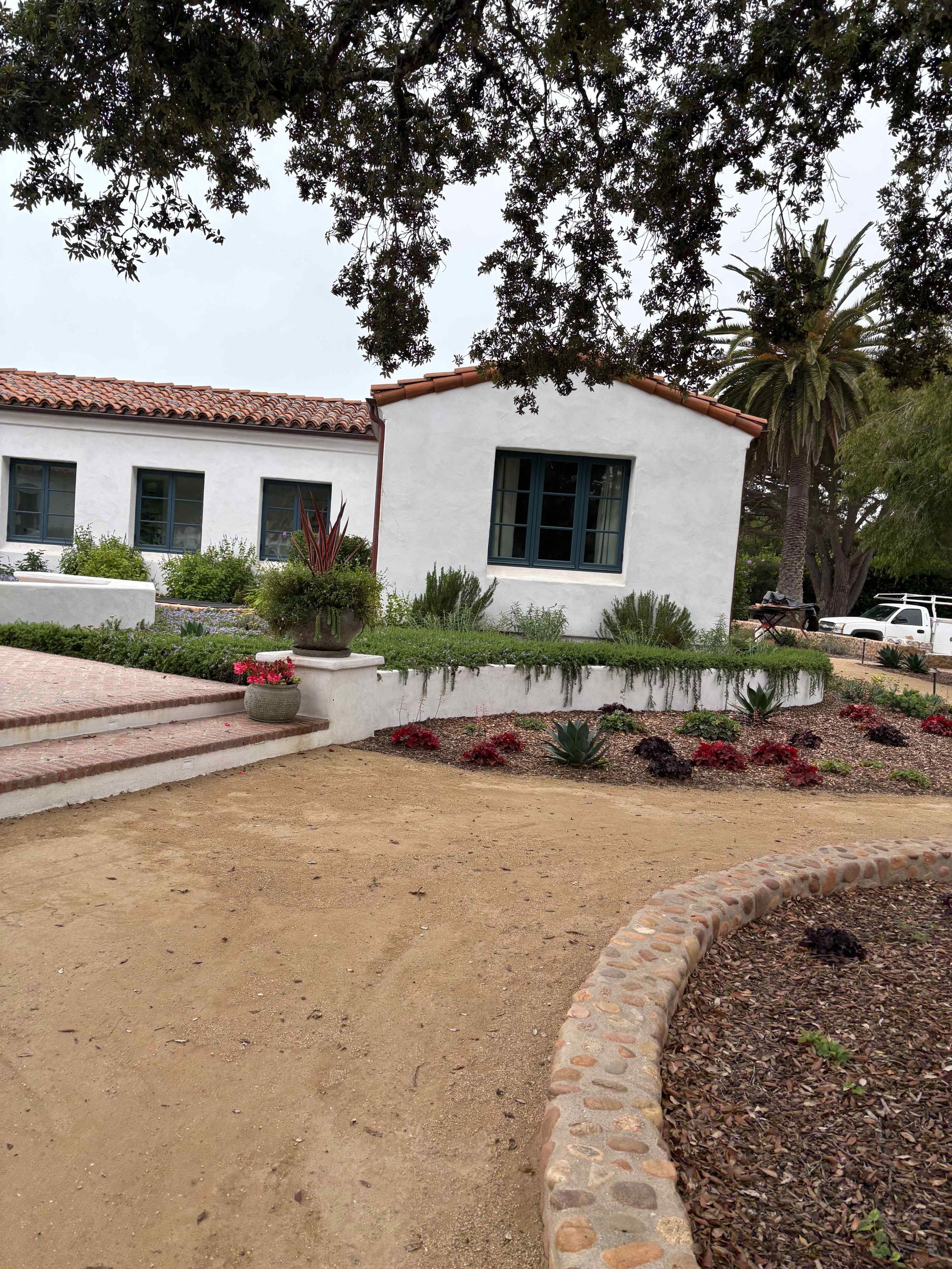 White stucco house with red tile roof, front garden, and curved gravel driveway under trees