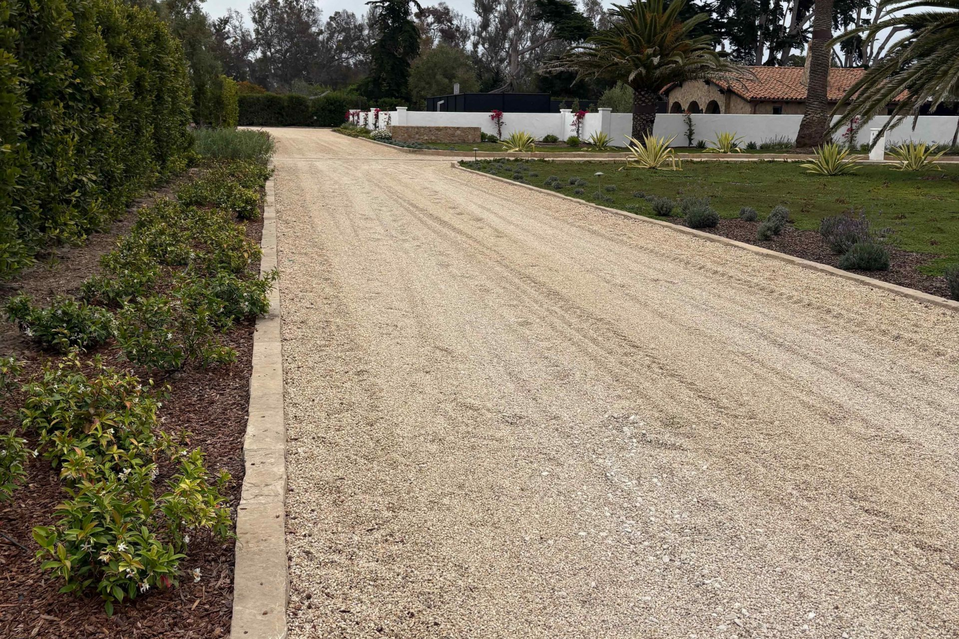 Gravel driveway beside hedges and garden beds leading to a white wall and trees