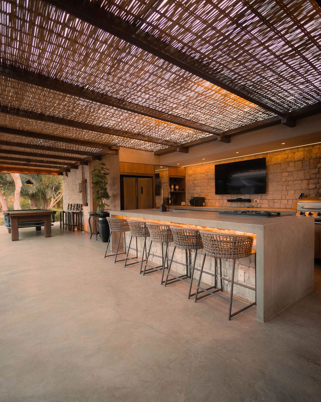 Open-air patio bar with stone counter, metal stools, woven ceiling, and TV on the wall