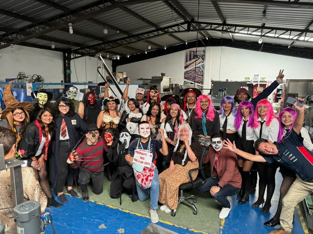 Un grupo numeroso posa en el interior de un espacio que parece un almacén, disfrazados para Halloween, sonriendo y levantando las manos.