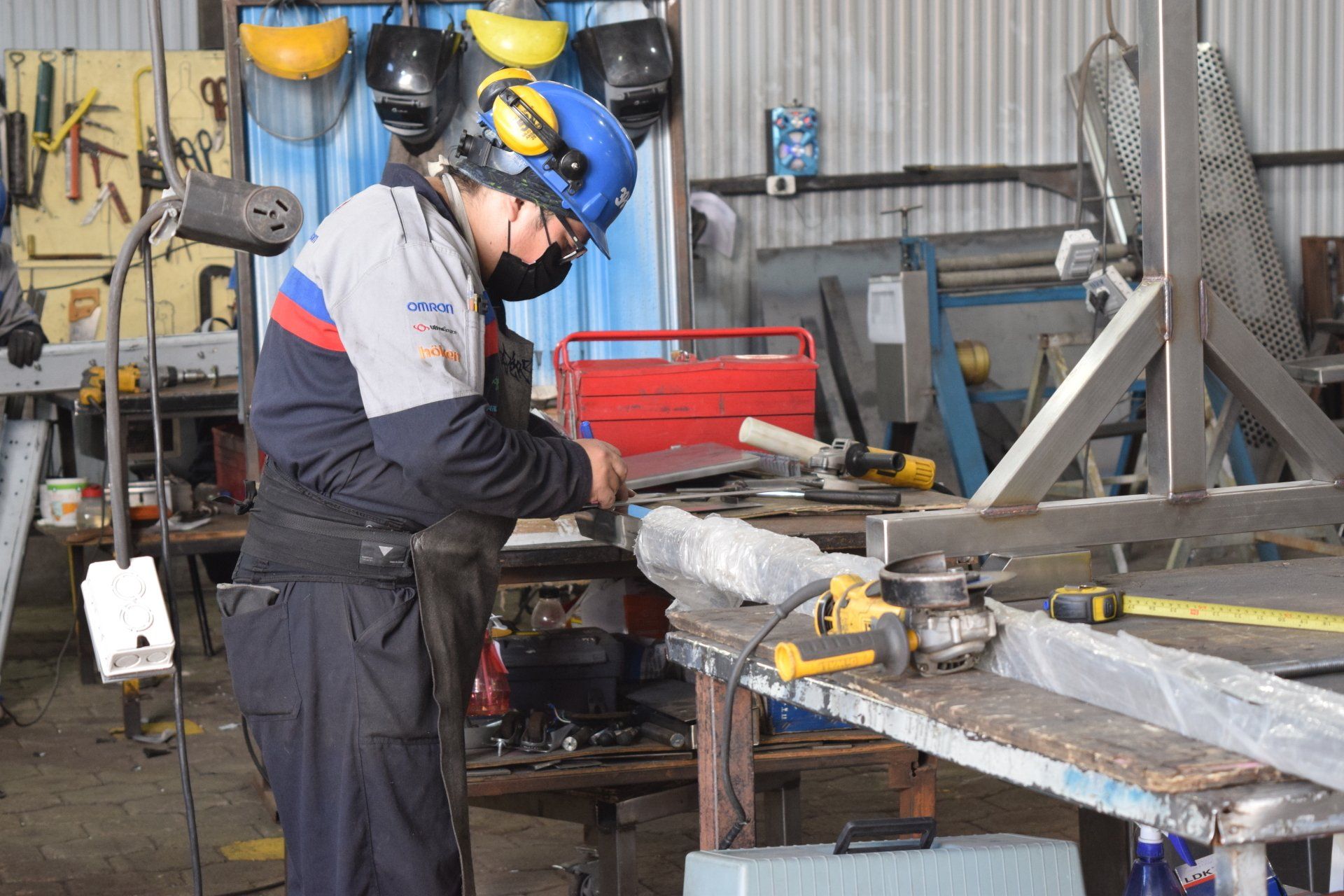 Un trabajador con casco azul soldando metal en un taller junto a una caja de herramientas roja.
