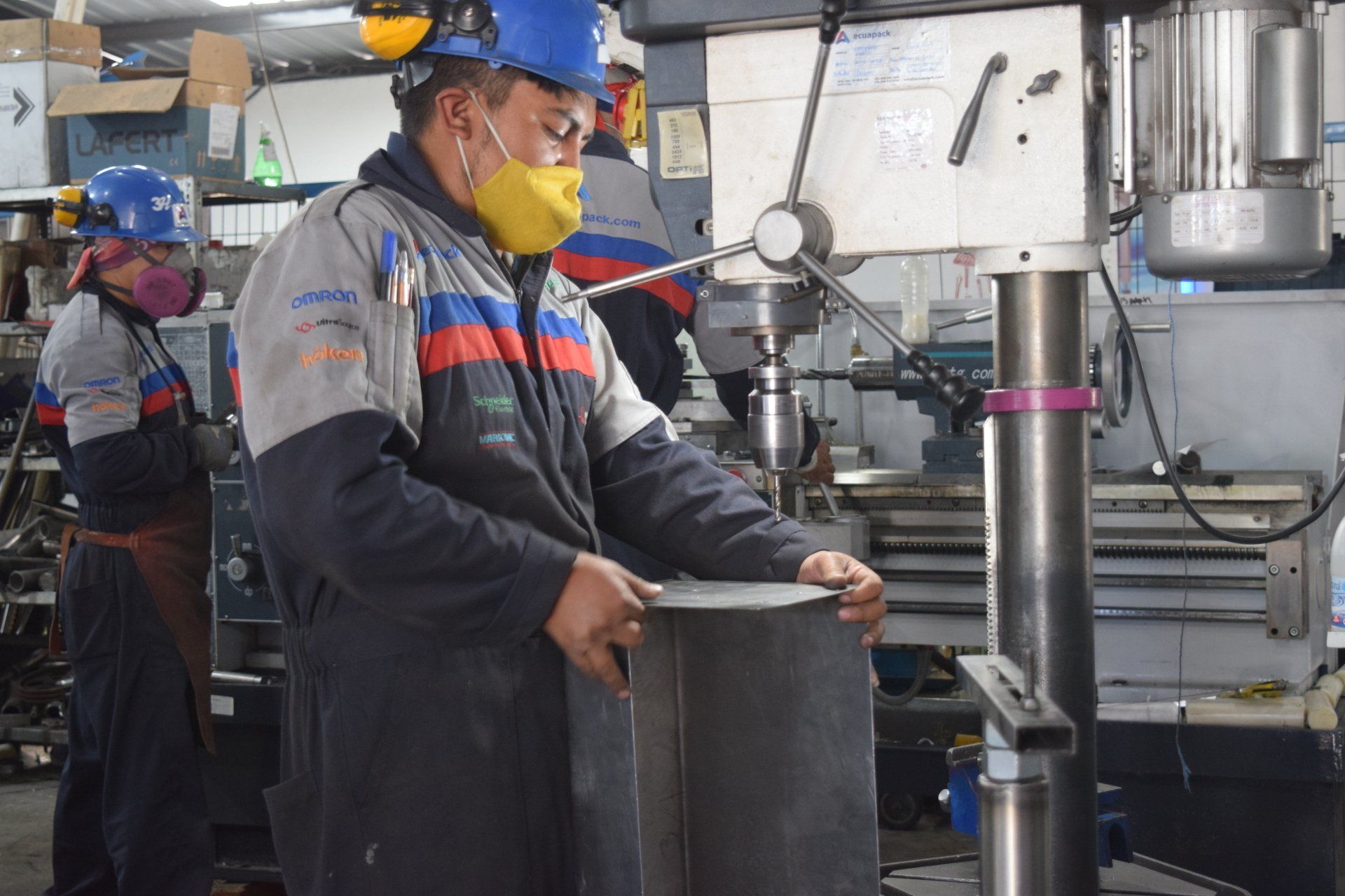 Obrero con casco y mascarilla manejando un taladro en un taller de fábrica.