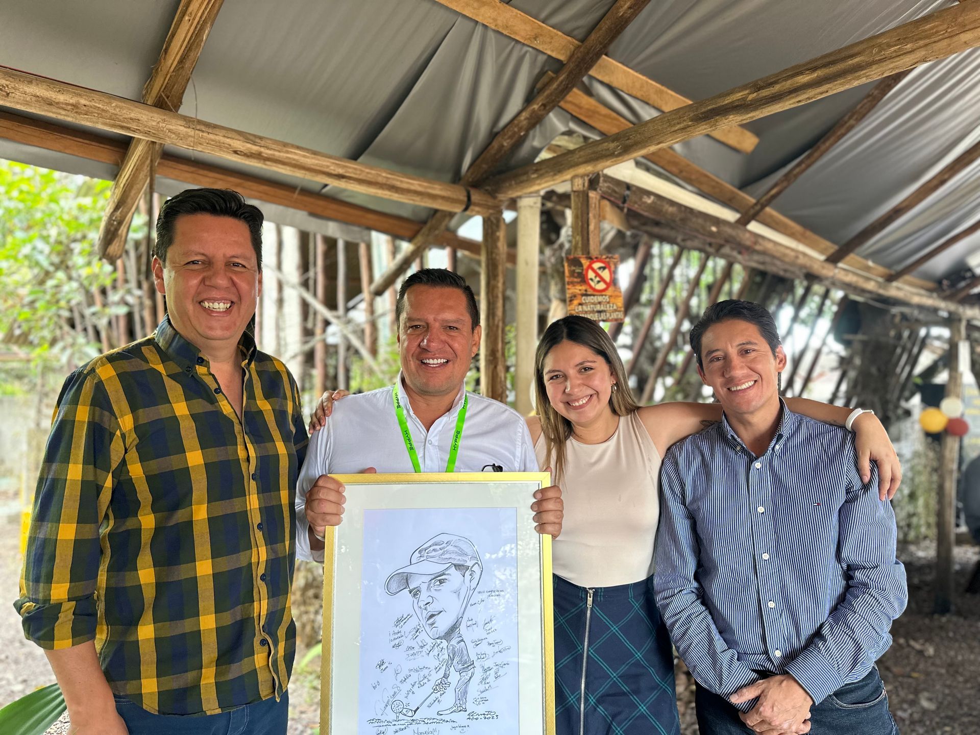Cuatro personas sonriendo en un refugio rústico, sosteniendo un boceto enmarcado.