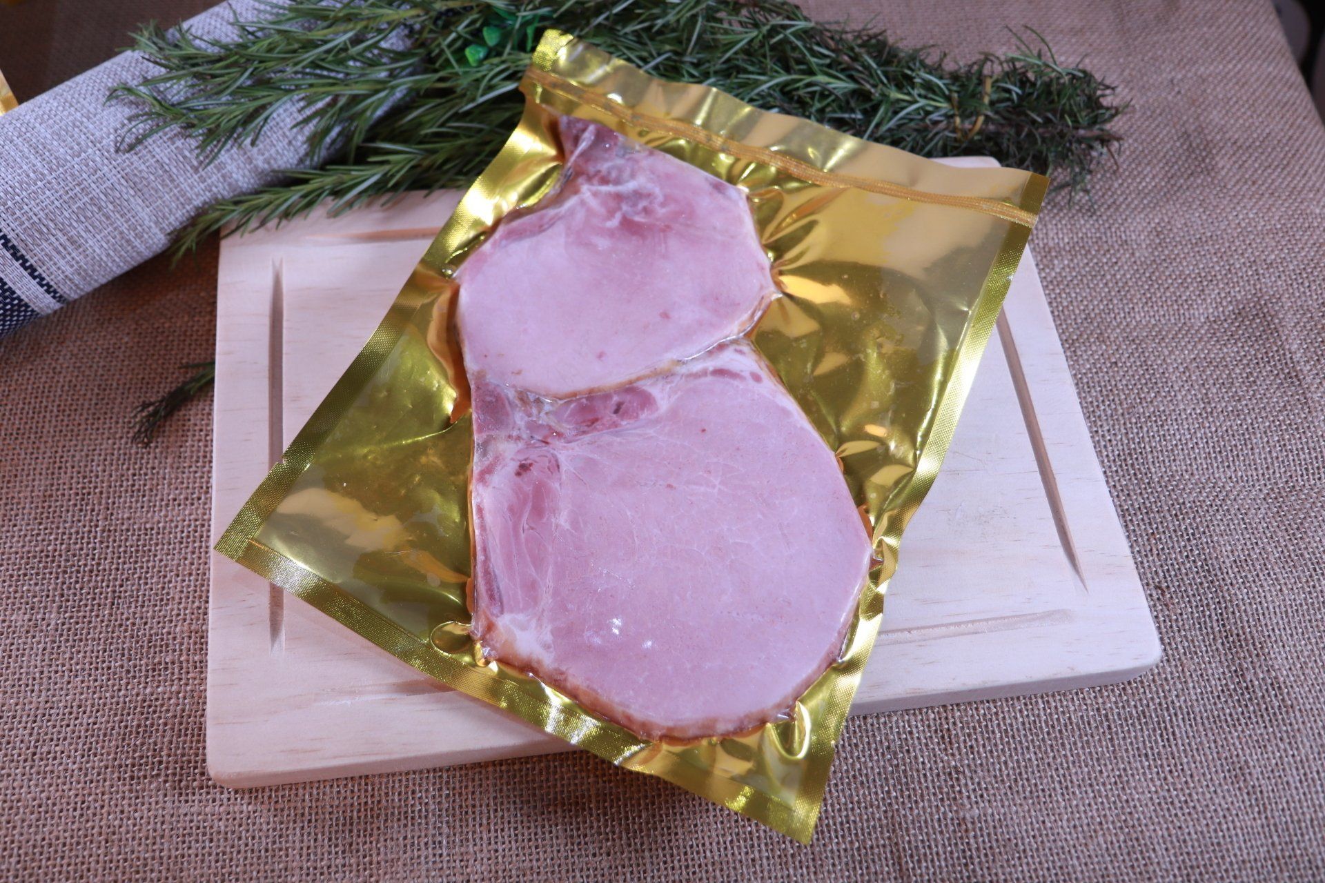 Dos lonchas de jamón rosado envueltas en papel dorado sobre una tabla de madera con vegetación al fondo.