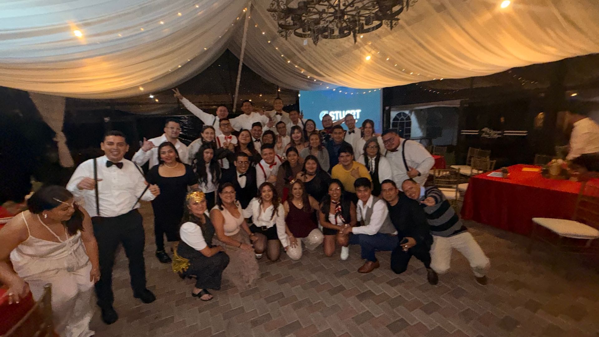 Un grupo numeroso posa para una foto bajo una carpa cubierta con cortinas en un evento festivo en interiores, con mesas e iluminación cálida.