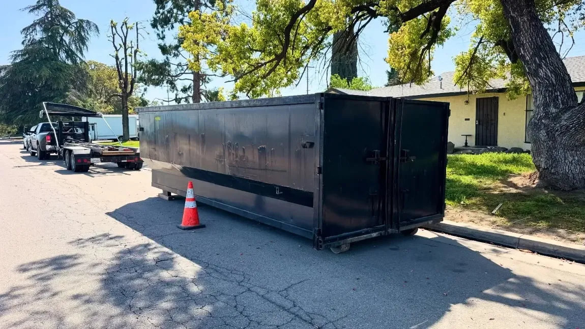 Black dumpster on wheels in street, orange cone in front. Tow truck in the background.