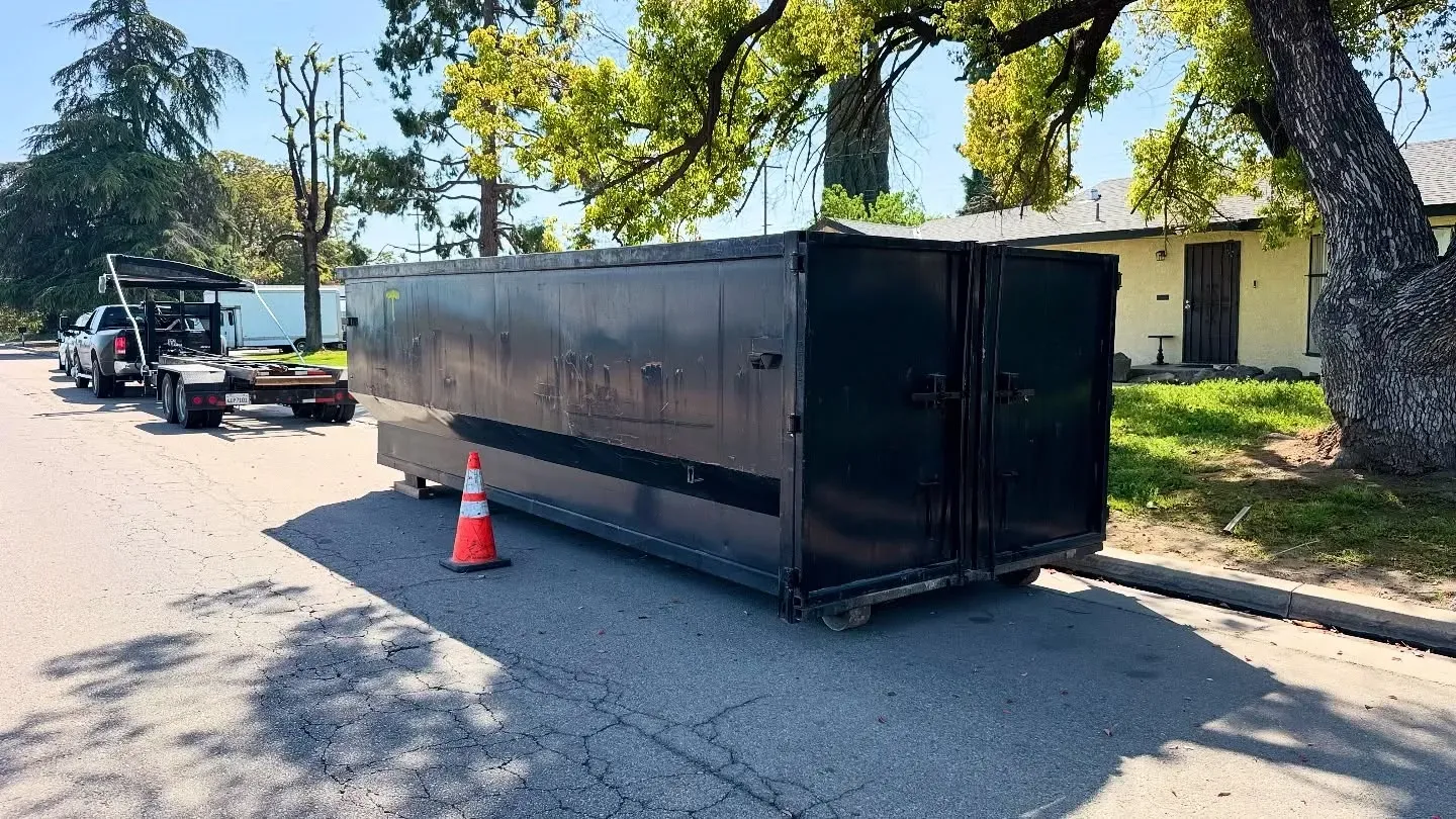 Black dumpster on wheels blocking a street, near a tree and house; tow truck in background.
