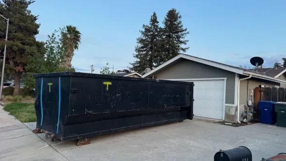 Black dumpster in driveway next to a gray garage, daytime, blue sky, trees.