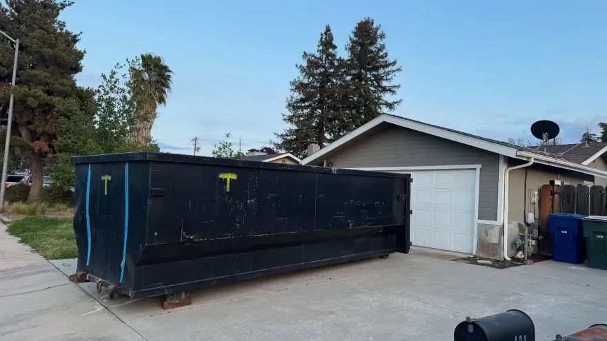 Black dumpster in driveway next to a gray garage, daytime, blue sky, trees.