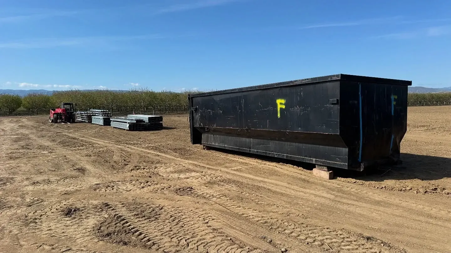 Black dumpster on dirt field; small tractor and stacked pallets in the distance. Sunny day.