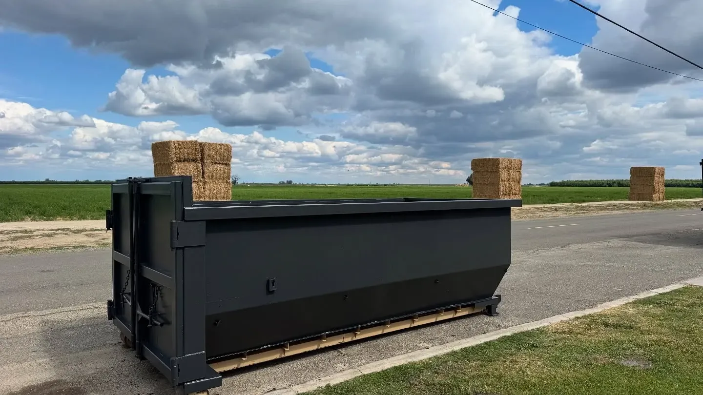 Black dumpster on a road in front of a green field and blue sky with clouds.